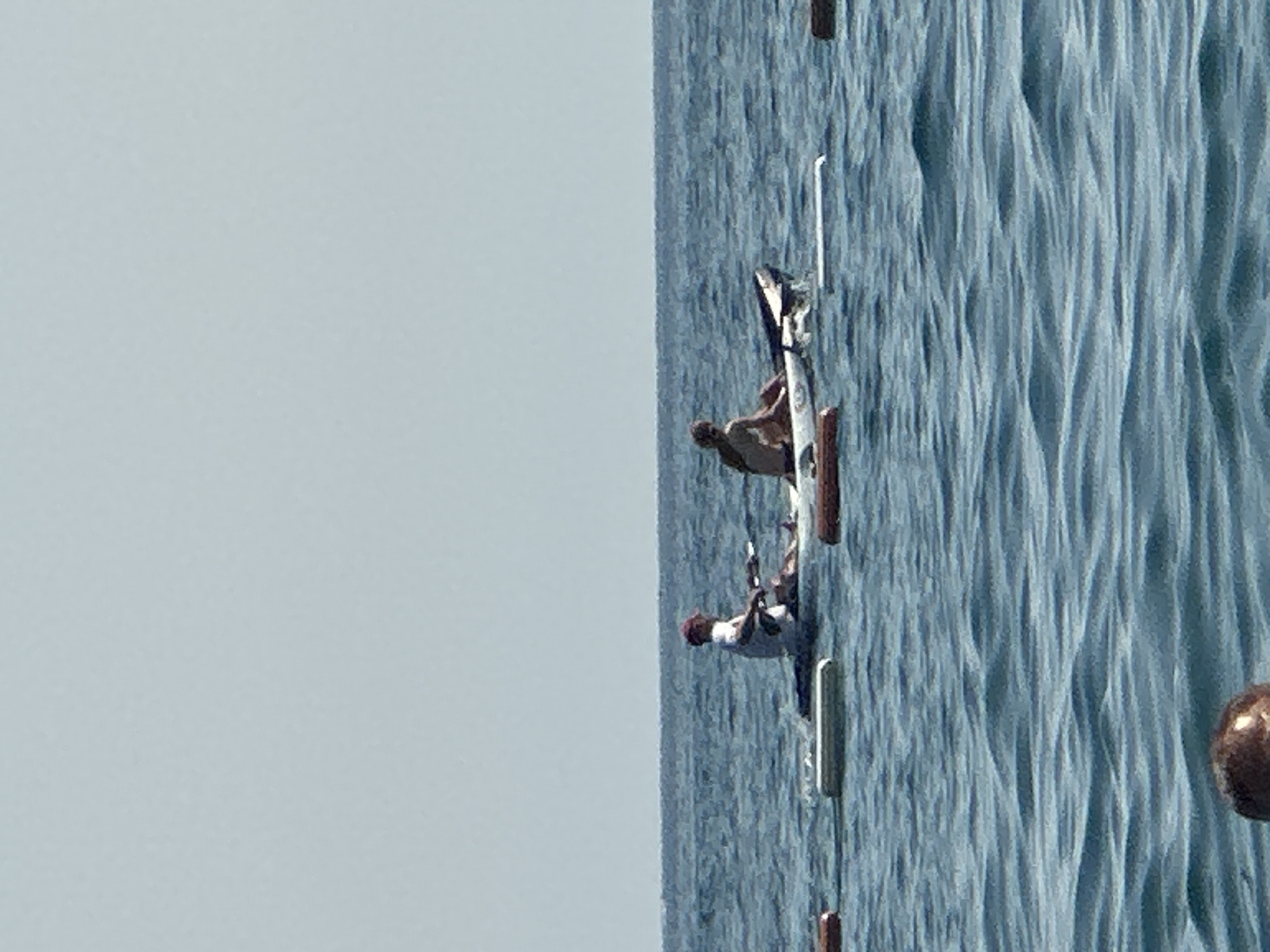 Two people are on a tandem kayak in the sea. The person in the front is wearing a white shirt and a red hat and is holding a paddle. The person in the back is wearing a light-colored swimsuit. The kayak is white with black accents. There are buoys in the water around the kayak. The sky is clear and pale blue. The water is calm with small waves. The scene appears to be at a beach in Boreti, Montenegro.