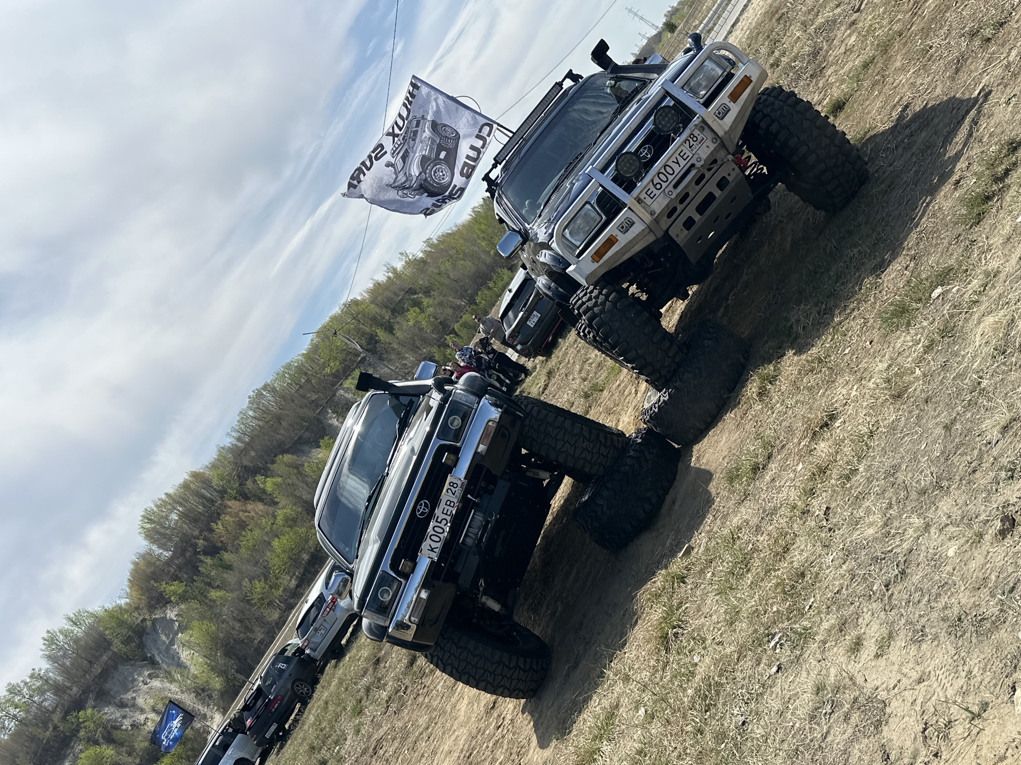 The image shows a gathering of off-road vehicles, primarily Toyota Hilux models, in a dirt/grassy area. License plates indicate the location as Russia. A "HILUX SURF CLUB" flag is attached to one vehicle. The vehicles are parked in a line, with a wooded area and a rocky outcrop visible in the background. The location could be near Sadovoe, Russia, but there is no definitive information to determine the exact location. The image does not contain information about any people, actions, or interactions.