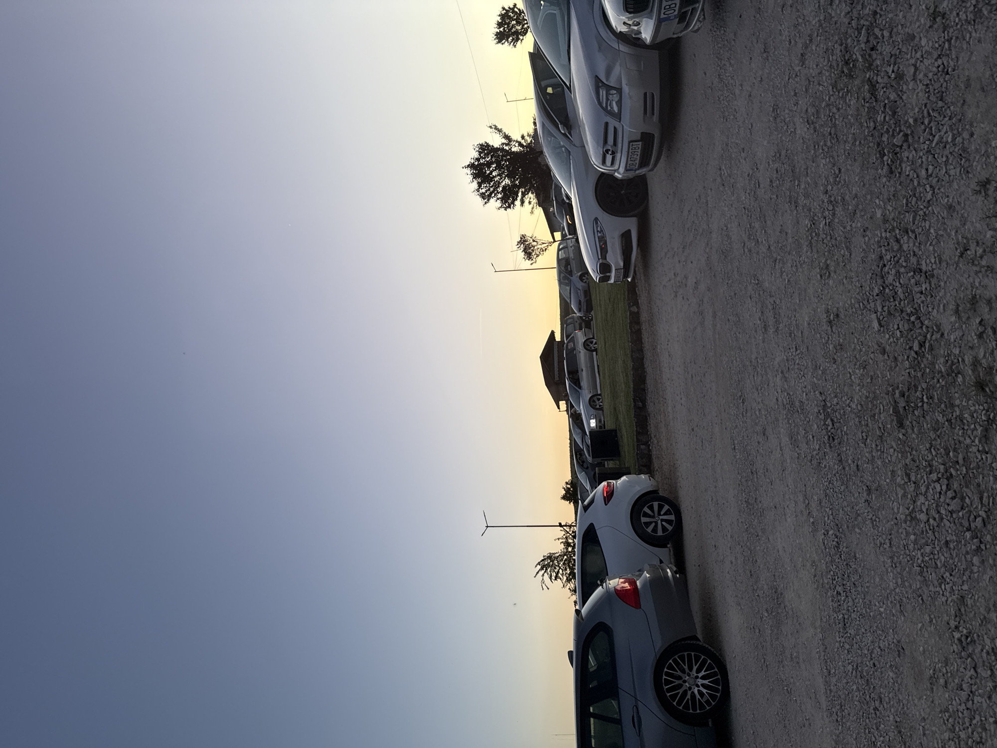 The image shows a line of parked cars in a lot. The location is likely Lukovit, Bulgaria. The sky is a gradient of blue to orange, suggesting dawn or dusk. There is a wind turbine visible in the background. The ground is gravel. No people are visible.