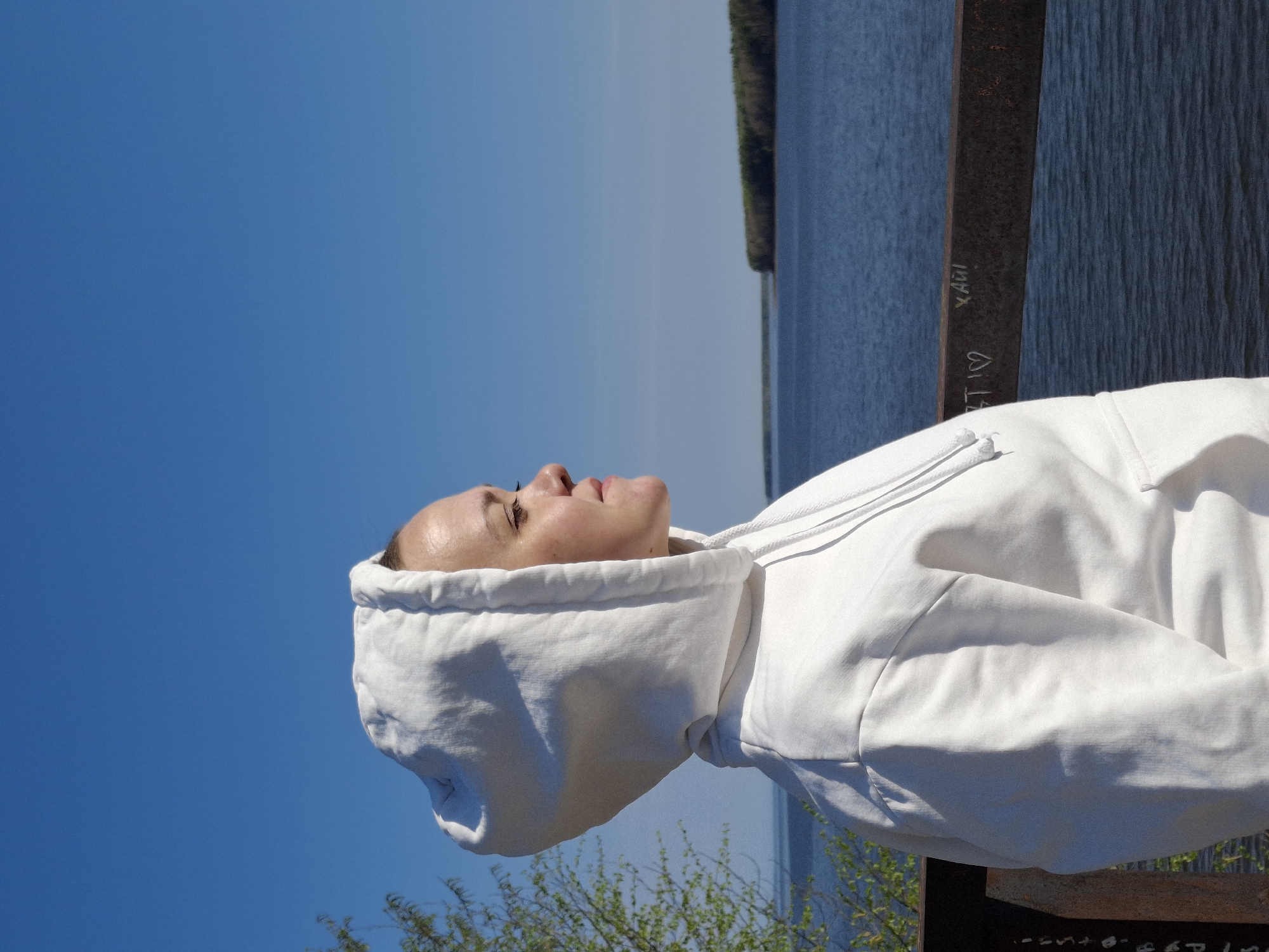 The image depicts a person wearing a white hooded sweatshirt, viewed from a low angle. The person is positioned to the right of the frame, with their face visible in profile, looking upwards. The background consists of a clear blue sky above, and a body of water in the upper right. The body of water is lined with trees. A metal structure with graffiti is visible, with the writing "X AUI" in white. The person appears to be outside, possibly on a bridge or elevated structure overlooking the water. The location is Chaikovsky, Russia.