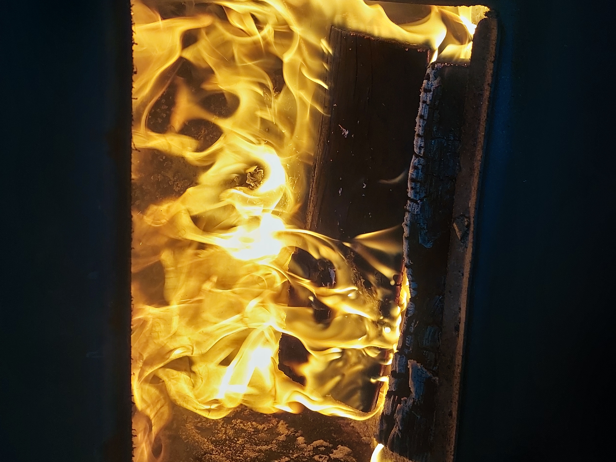 The image shows a close-up view inside a wood-burning stove or fireplace. Two pieces of wood are visible, with flames enveloping them. The wood on the right appears charred. The surrounding area is dark, suggesting the stove is enclosed. The location is not discernible from the image itself. The image contains no people, actions, or interactions.
