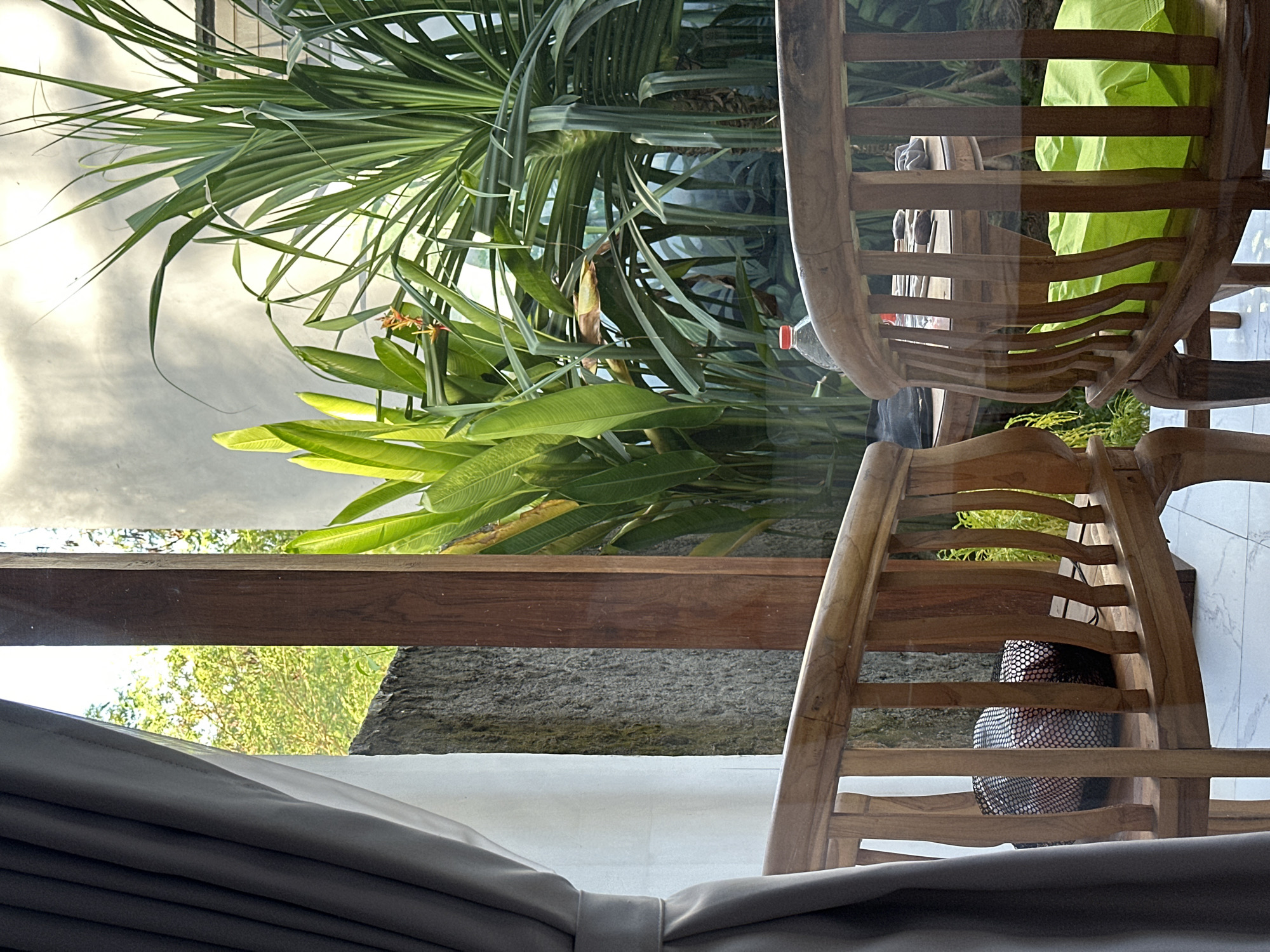 The image depicts an indoor scene with a view to the outside featuring lush greenery and palm trees, likely located in Nusa Penida, Indonesia, given the tropical foliage. Two wooden chairs are partially visible in the foreground, suggesting a relaxed setting. The view through the window includes a partial view of the sky and possibly other buildings, indicative of a semi-urban environment. The presence of drawn curtains hints at an interior space designed for privacy or shade from the sun.