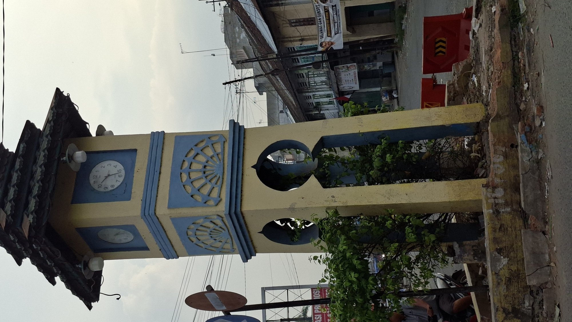 The image shows a faded yellow and blue clock tower in Tanjung Pura, Indonesia. The clock tower has a tiered roof with dark shingles and multiple lights. One clock face is clearly visible, showing the time as approximately 10:50. The tower has decorative blue fan-shaped elements and arched openings. Bushes and trees grow around the base of the tower. In the background, there are buildings with weathered facades, some with signs and banners, suggesting a commercial area. Electrical wires crisscross the sky. Red traffic barriers are visible on the right side of the road. A person is partially visible on a motorcycle on the left side of the image. The overall scene depicts an outdoor, urban environment with signs of age and wear.