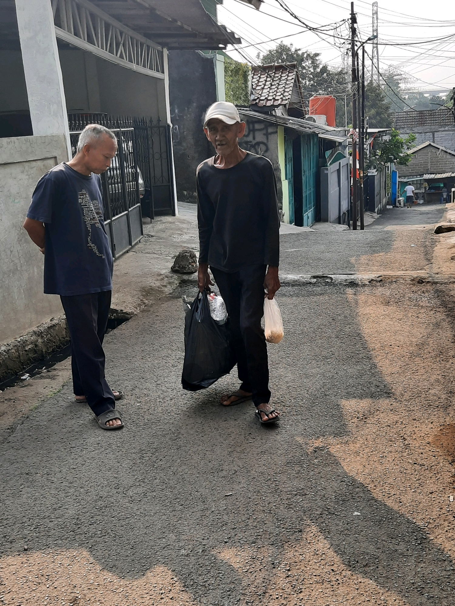 Two elderly Asian men are standing on a paved alleyway in a residential area of East Jakarta, Indonesia. The man on the left, with grey hair, is wearing a dark blue t-shirt with white text and dark pants. He is facing away from the camera and looking down. The man on the right, wearing a white baseball cap, is wearing a black long-sleeved shirt and dark pants. He is holding a black plastic bag in his left hand, containing what appears to be bottles of water, and a small white plastic bag in his right hand, containing food. The alleyway is lined with modest houses and buildings. Overhead, a complex network of electrical wires is visible. The ground surface is asphalt with some patches of water and debris. The lighting suggests it is daytime, with shadows cast across the alley. In the background, more houses and a water tower are visible, along with distant power lines and structures.