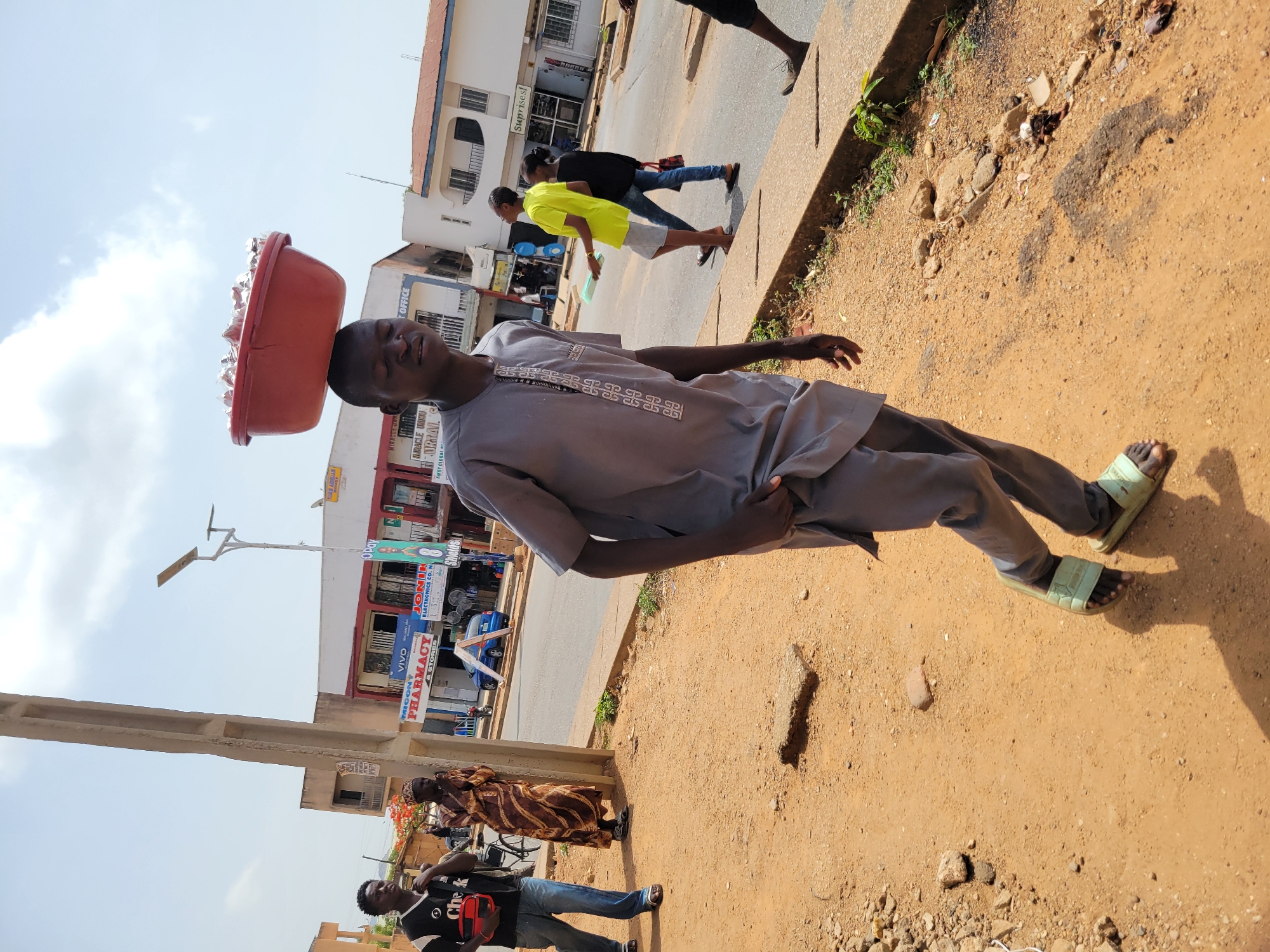 A man balances a red basin on his head while walking on a dirt shoulder next to a street in Jos, Nigeria. The basin appears to contain smaller packages. The man is wearing a gray, short-sleeved shirt and pants, with light green sandals. He has his left hand in his pocket and his right arm extended. In the background, people are walking on the sidewalk and across the street. Buildings with commercial signage, including “VIVO Pharmacy & Stores” and “JONIK Electronics Co. N”, line the street. A light pole is also visible. The ground is dusty and littered with small rocks.