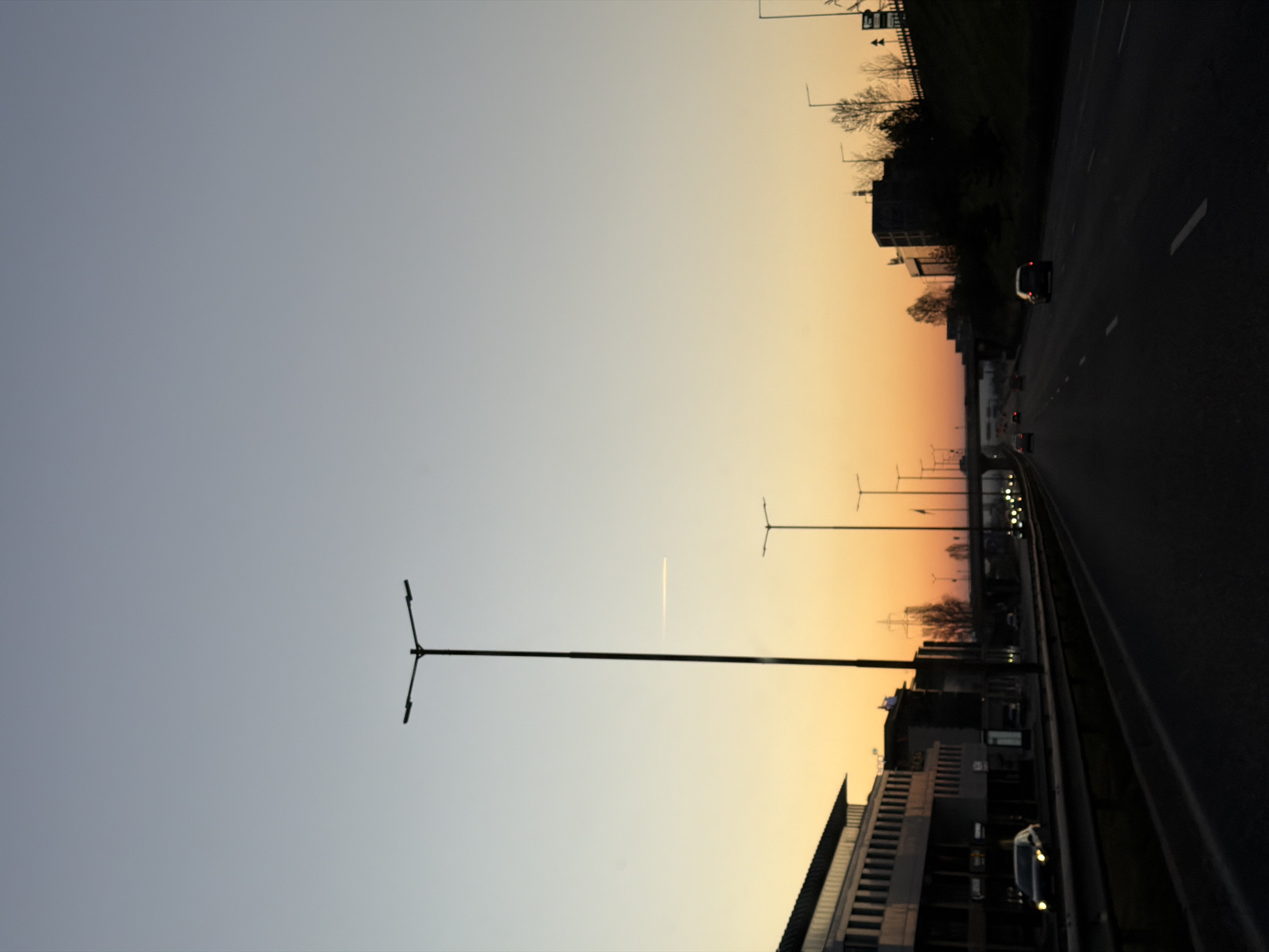 The image depicts a highway in Sofia, Bulgaria, during sunset. The sky transitions from grey to orange. Streetlights line the highway. Buildings are on one side of the highway, and vegetation lines the other. Several cars are visible on the road.
