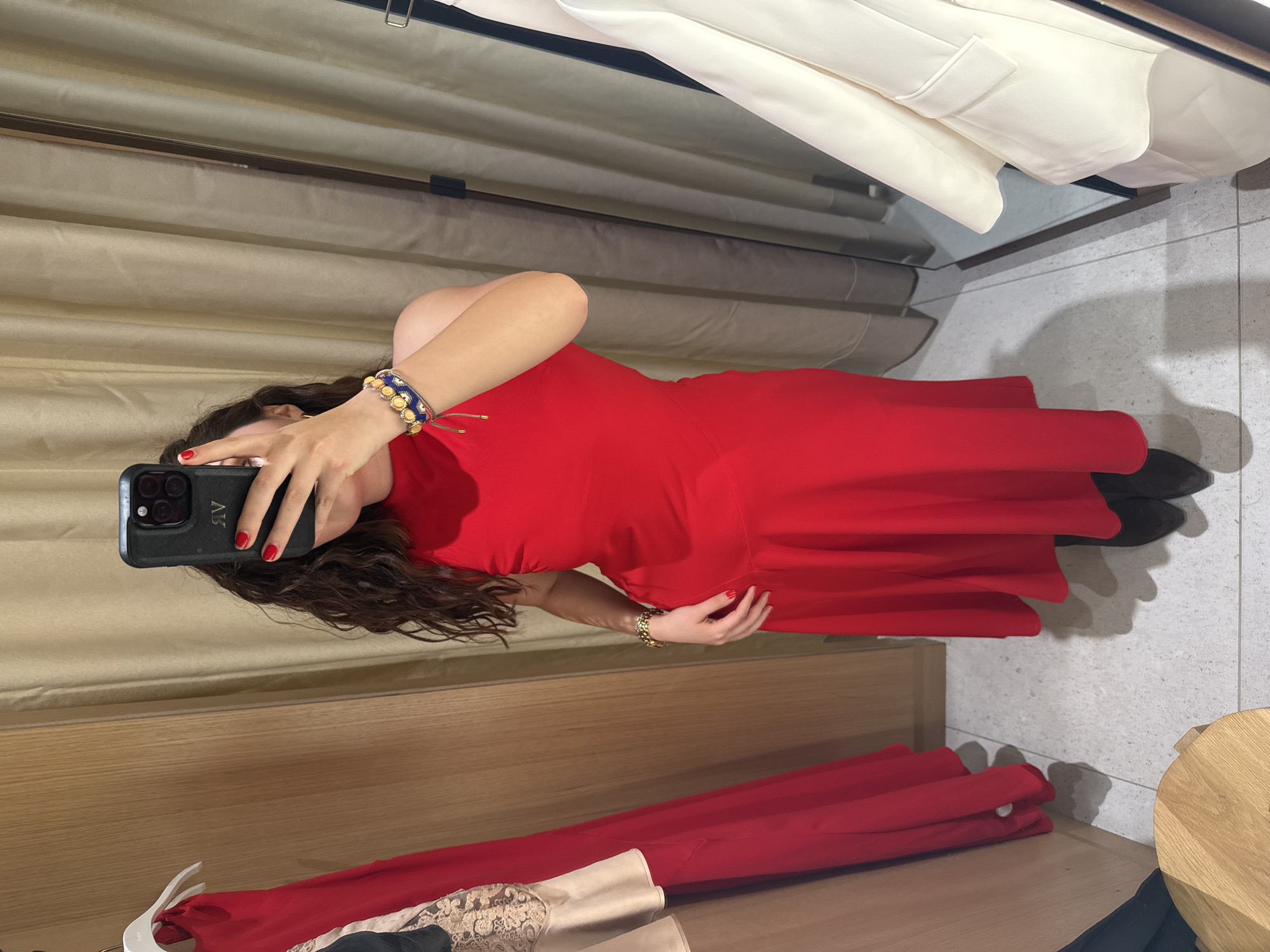 Here is a detailed description of the image:
The image appears to be a mirror selfie taken in what looks like a clothing store fitting room. The person taking the photo is wearing a bright red dress with a flared skirt, and dark shoes. A bracelet is visible on the wrist that is holding the phone, and another bracelet is on the other wrist. The phone case is dark, and the nails are painted red.
The fitting room has a beige curtain as the backdrop and a wooden bench or surface on which other pieces of clothing are placed. These garments are a mix of red, beige, and what looks like lace material. Above, there's a rack of white clothing, possibly coats or blazers. The floor is tiled.
The location is potentially in Mexico City, Mexico, given the query request. However, without further context or specific details unique to the city, it is hard to confirm the location definitively.