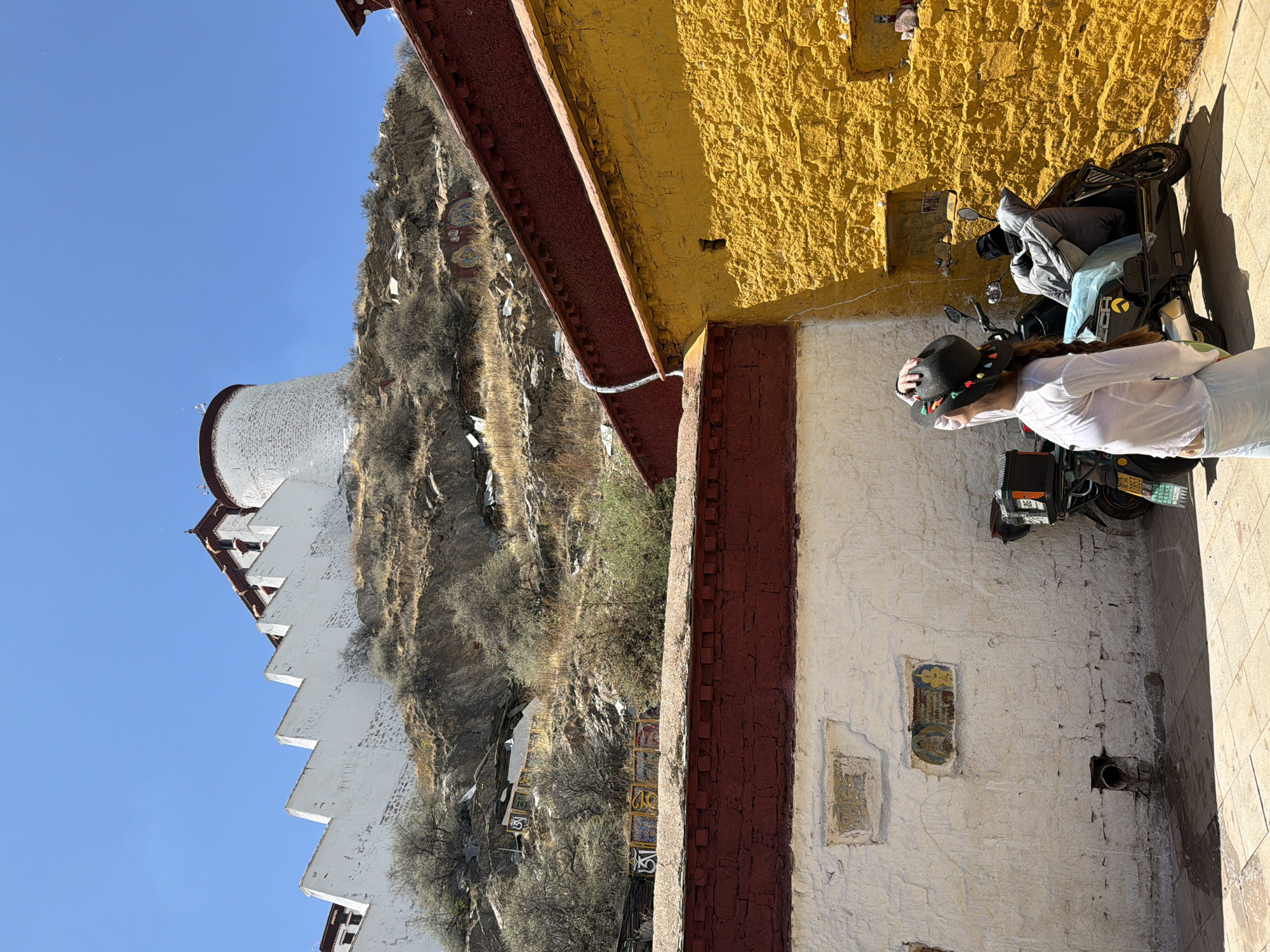 The image depicts a bright, sunny outdoor scene in Chengguan District, China, showcasing distinctive architecture and natural landscape. The main subject is a person standing near a black scooter. The individual, seen from behind and side, wears a white long-sleeved shirt, light-colored pants, and a black straw hat decorated with colorful tassels. They appear to be observing the scene to their right.
The setting features traditional Tibetan-style architecture. In the foreground, there's a building with a textured bright yellow upper wall and a white lower wall, separated by prominent dark red beams and columns. Both wall sections have recessed areas with faint, colorful painted artwork. The ground is paved, and strong sunlight suggests it is midday with a clear blue sky.
Behind the foreground building, a steep, rocky hillside rises, covered in sparse, dry vegetation. Large, colorful painted or carved symbols, resembling Tibetan mantras like "Om," are visible on the hillside, along with numerous white square prayer flags or offerings. Dominating the left side of the scene is a majestic multi-story white building, possibly part of a monastery or palace complex, featuring stepped tiers, many windows, and a distinctive round, white-domed top, resembling a stupa, against the clear blue sky.
The black scooter parked beside the person has a license plate visible at the rear, bearing the characters "西藏" (Xizang, meaning Tibet) followed by "A" and some numbers/letters, partially obscured. The overall atmosphere is peaceful and culturally rich.
