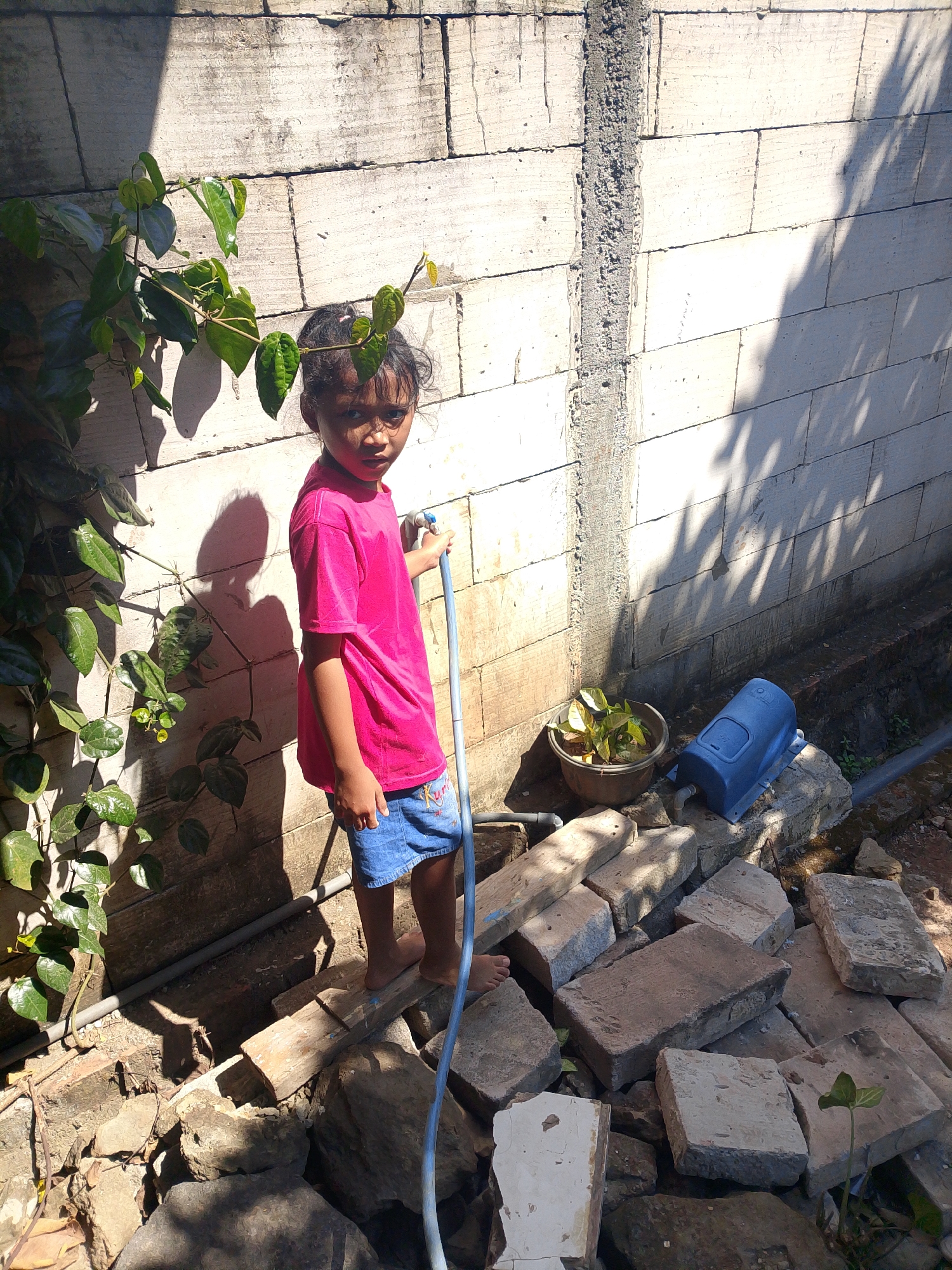 In the city of Penyangkringan, Indonesia, a young girl stands on a wooden plank, holding a blue hose. She is wearing a bright pink t-shirt and denim shorts. The background is a rough-textured, light-colored brick wall, partially covered by the shadow of leafy branches from a nearby plant. The ground is strewn with various debris, including bricks, stones, and a small potted plant. A blue plastic object, possibly an aerator, sits on a stone to the right of the potted plant. The scene is bathed in bright sunlight, casting sharp shadows.