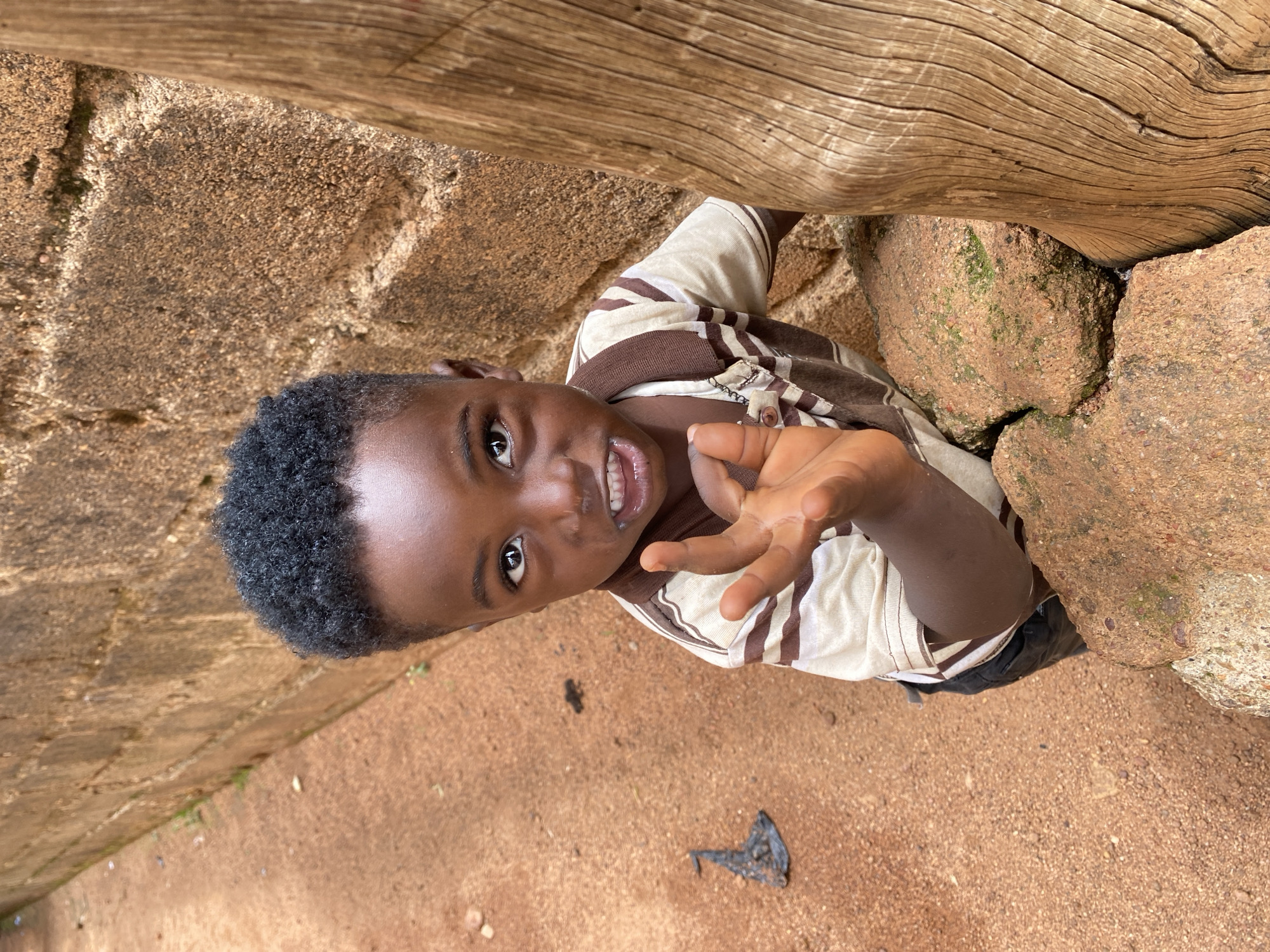 The image, taken in Kuru Dungus, Nigeria, captures a cheerful young boy in an outdoor, natural setting. The boy, appearing to be of African descent, is the central subject, viewed from an overhead perspective looking up at the camera. He has short, dark, curly hair and a bright smile, revealing his teeth, with wide, engaged eyes. He is wearing a light-colored t-shirt with brown stripes and dark-colored shorts or trousers. His right hand is raised towards the camera, fingers slightly splayed, in what appears to be a friendly gesture or greeting.
The background features large, rough-textured reddish-brown rocks, some covered with patches of green moss, suggesting a natural and somewhat rustic environment. A weathered wooden log or beam with prominent grain runs horizontally across the upper part of the frame, positioned behind and above the boy. The ground is reddish-brown dirt or sand. A small, dark, crumpled piece of debris, possibly a leaf, is visible on the ground near the boy's lower right. The scene is illuminated by bright natural daylight, indicating daytime. There is no visible text in the image.