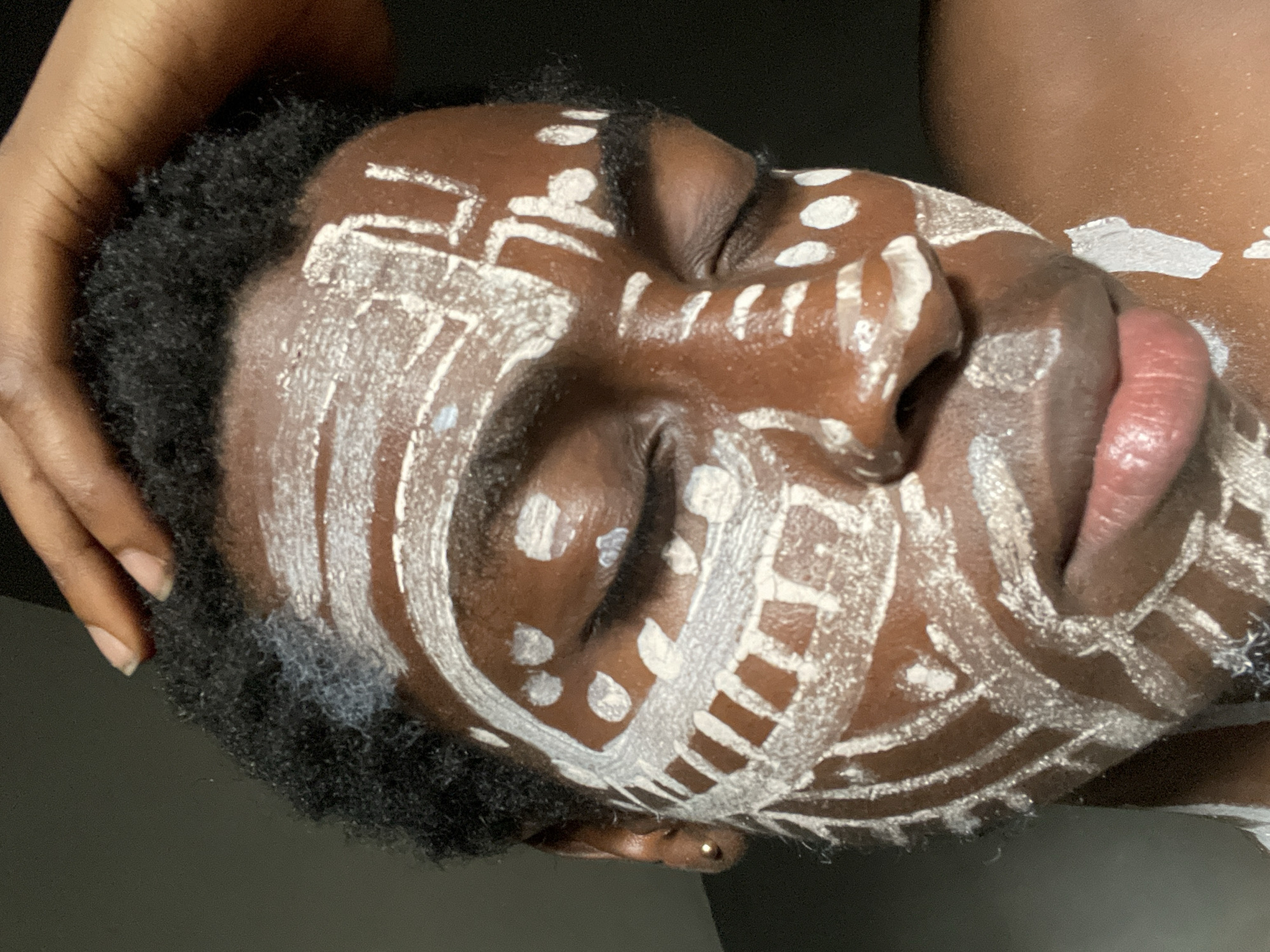 A close-up portrait showcases a person's head and upper body, intricately adorned with white body paint, set against a dark, out-of-focus background. The individual, appearing to be in Onitsha, Nigeria, has dark skin and short, kinky black hair that frames the face. The head is gently tilted upwards and to the side, with eyes closed or cast downwards, conveying an expression of serenity and contemplation.
The white body paint covers the forehead, cheeks, nose, and extends across the lips and down the neck and shoulder. The patterns are diverse, featuring bold parallel lines, zig-zags, and clusters of dots, applied with precision. The paint's texture varies, from thick and opaque patches to thinner applications that allow the underlying skin tone to subtly show through. The lips are slightly parted, revealing their natural pinkish hue. A small gold stud pierces the earlobe. In the upper left corner, a hand, likely of another person or the subject, is seen gently cradling the head. The lighting highlights the contours of the face and the intricate details of the body art, suggesting a cultural ritual, performance, or artistic expression. No visible text is present in the image.