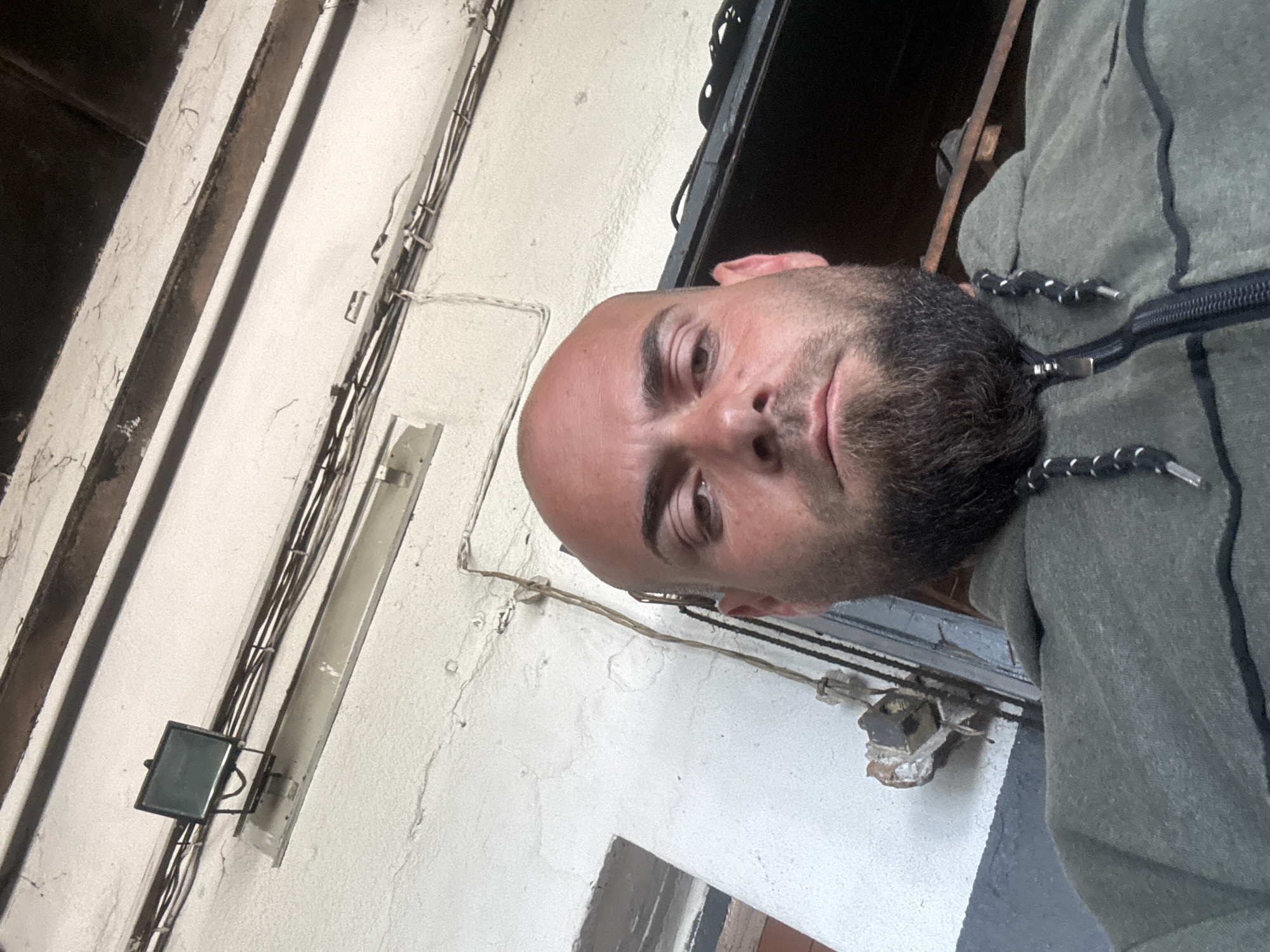 A man with a bald head and a dark beard is prominently featured in a close-up, upward-angled shot. He looks directly at the camera with a neutral expression. He is wearing a dark greenish-grey zippered hoodie with black trim and white-and-black striped drawstrings.
The background consists of a textured, light-colored, possibly off-white, wall that shows signs of age, including cracks and some discolored patches. Numerous electrical wires and cables, dark in color, are visibly routed along this wall, some bundled together. Various aged electrical fixtures and mountings are attached to the wall. To the man's left, a small, rectangular fixture with a dark screen or glass cover is visible, possibly a security camera or a light. Above it, a long, translucent, tube-like fixture, resembling an old fluorescent light cover, is also attached to the wall.
To the man's right, a dark, recessed area suggests a doorway, window, or an opening into another section of a building. The overall setting appears to be an older, functional, or utility-focused area, possibly an interior space or a covered outdoor passageway within a building in Sarnevo, Bulgaria. The lighting is diffused, suggesting either an overcast day or an artificially lit environment, with no strong shadows evident. There is no visible text.