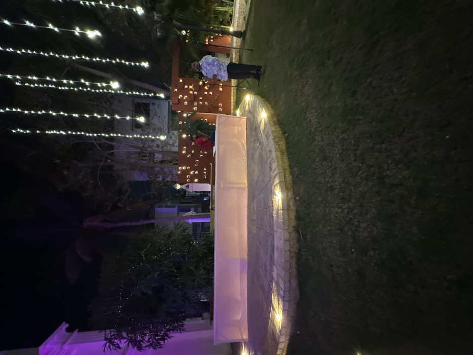 The image depicts a nighttime outdoor scene, consistent with an event or celebration setting. The specific location within Vadodara, India, is not discernible from the visual information provided.
In the foreground, a dark green grass lawn extends across the lower portion of the frame. A semi-circular raised platform, paved with light-colored stone tiles and bordered by brick, is positioned centrally. Small, embedded warm-toned spotlights illuminate the edge of this platform.
In the midground, on the raised platform, a long, rectangular structure, appearing to be a stage or table, is covered with a translucent light-colored fabric, illuminated from within or beneath with a soft glow. Behind this structure, slightly to the right, a male individual in a red shirt is partially visible, seated or crouching behind a small green plant. To his right, a decorative reddish-brown wall or partition features multiple square and rectangular cutouts through which warm light emanates. Standing on the grass to the right of the platform, another male individual wears a light-colored, patterned shirt and dark trousers, facing towards the left. He holds a dark, thin object vertically in front of him.
The background features numerous strings of small, warm white fairy lights hanging vertically, creating a canopy effect. Trees and dense foliage are visible behind the decorative wall and stage area, with some foliage also adorned with small lights. A portion of a light-colored building with architectural details, possibly illuminated with a purple uplight, is visible on the far left. The sky is dark, indicating night.