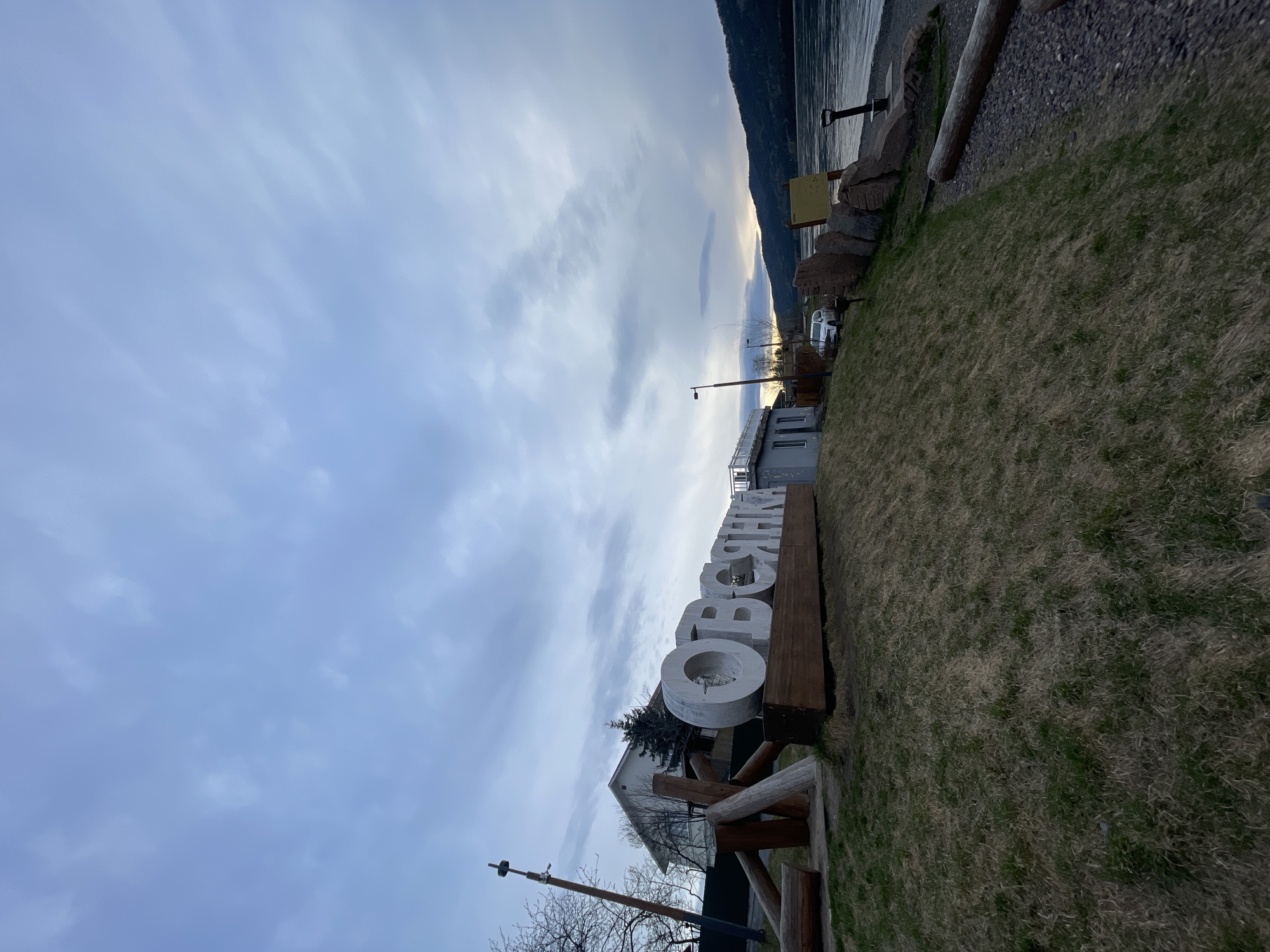 The image depicts a landscape scene featuring the word "ОВСЯНКА" spelled out in large, white, three-dimensional letters, suggesting a location in Овсянка, Russia. The letters are aligned horizontally on a wooden base. Behind the sign is a building with white walls and a gray roof, partially obscured by the letters. A street lamp is visible near the building. The foreground consists of grassy ground, partially covered with dry vegetation. In the background, there is a body of water with an elevated shoreline, potentially a river or a lake. The sky is overcast with varying shades of blue and gray clouds.
