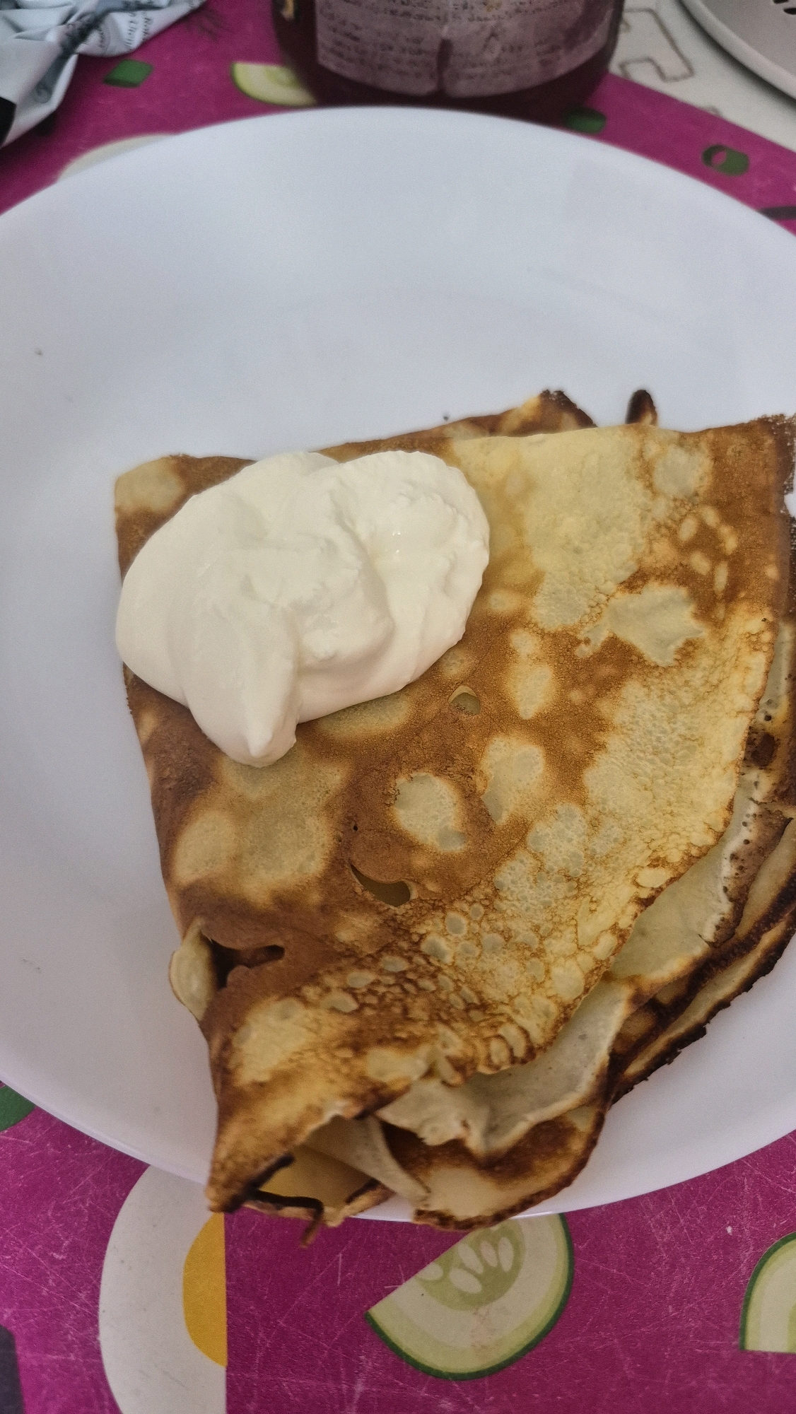 A close-up, slightly angled shot captures a plate of crepes topped with a dollop of cream. The crepes are golden brown with some darker, slightly crispy edges, indicating they've been cooked on a griddle. The cream is a bright white, smooth, and appears to be a thick sour cream or creme fraiche, piled high on the top crepe. The white plate is visible beneath the crepes, and the background is a colorful, patterned tablecloth with shades of pink, green, and white, suggesting a home or casual dining setting. The overall impression is of a simple, delicious dessert or breakfast.