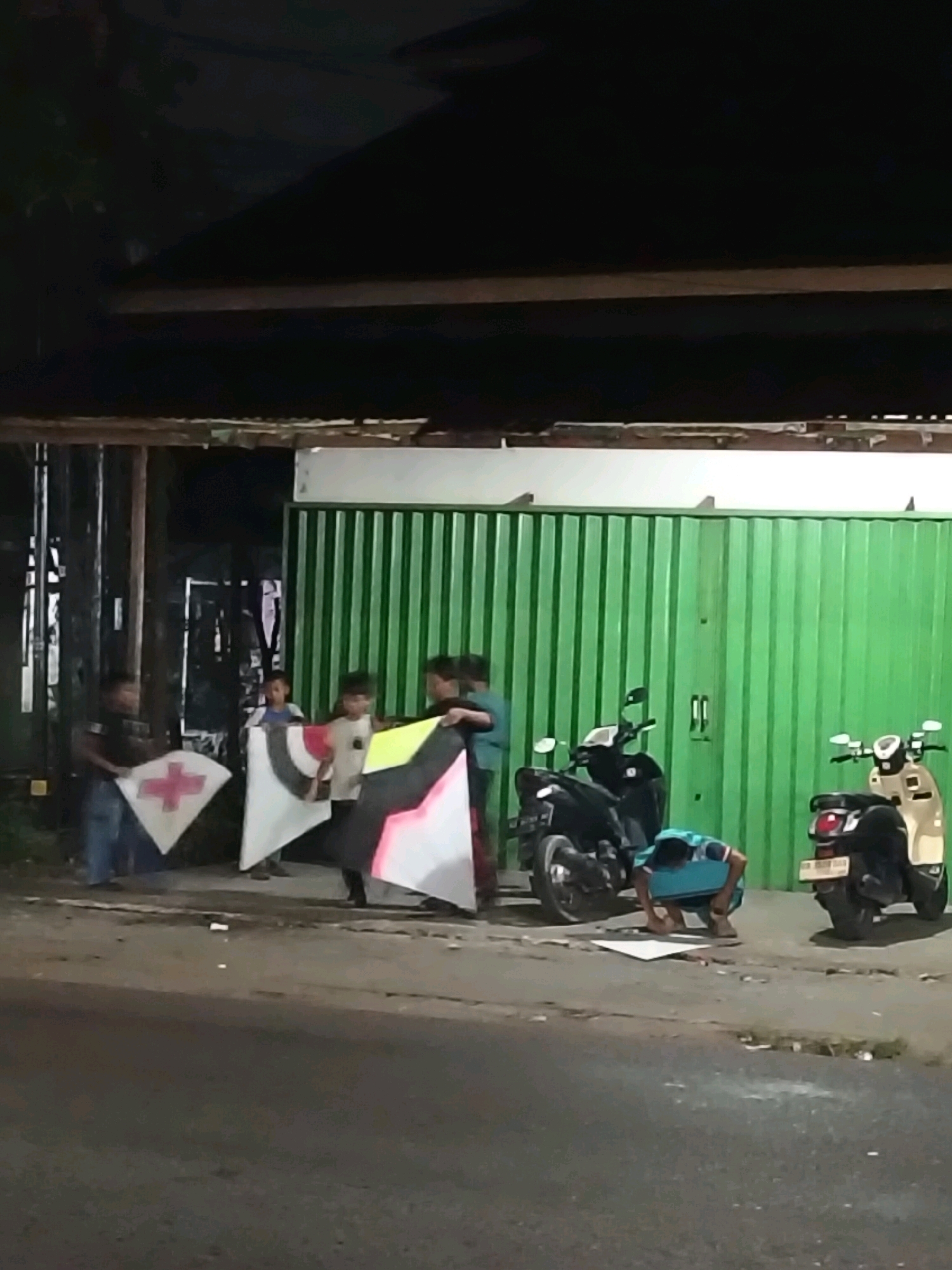 In Arang Limbung, Indonesia, at night, a group of individuals are gathered near a green corrugated metal shuttered building. In the foreground, a paved road is visible, with debris scattered on its surface. To the right of the image, two motorcycles are parked; a black one and a lighter colored one. A person is crouched on the ground in front of the black motorcycle, appearing to be working on something.
To the left of the building, several people are standing and holding large, flat objects, which appear to be kites. One kite is white with a red cross design. Another kite is white with black and pink geometric patterns. The figures are illuminated by artificial light, casting shadows on the ground. The overall scene suggests an outdoor gathering or activity taking place after dark.