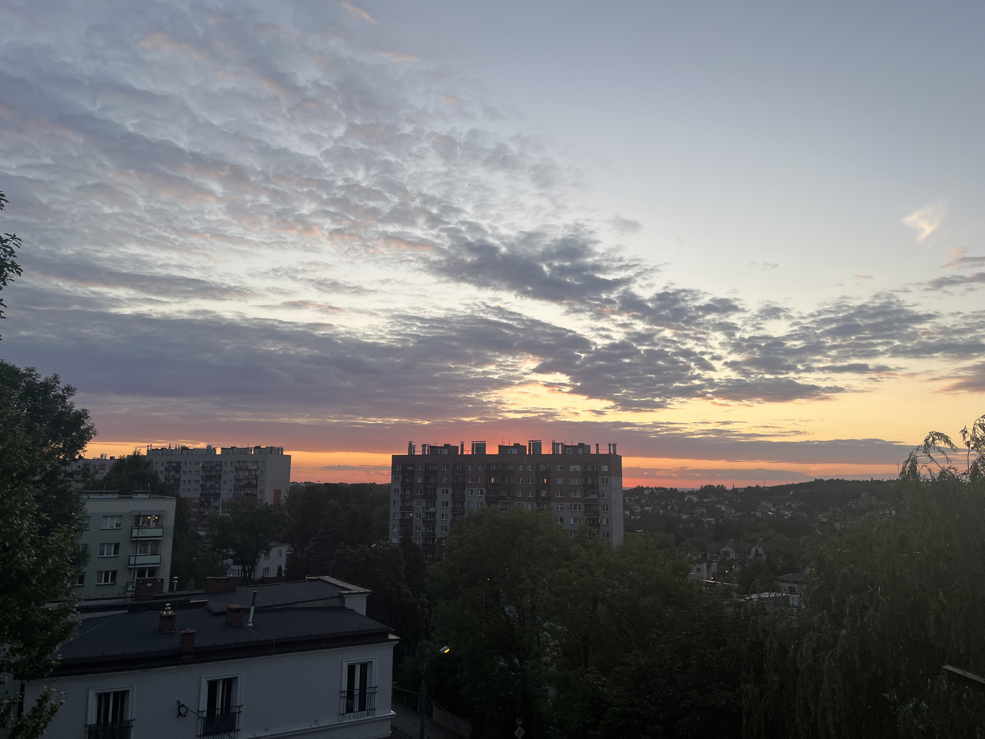 An outdoor, high-angle, eye-level medium shot captures a city skyline at dusk. The sky is a gradient of soft colors, transitioning from a pale blue at the top to a vibrant orange and pink near the horizon, indicative of a sunset. Scattered across the sky are numerous clouds, some appearing wispy and light, others darker and more textured, tinged with the colors of the sunset.
Dominating the middle ground of the image are several apartment buildings. The most prominent is a tall, multi-story building with a distinctive patterned facade and a flat roof with visible structural elements. To its left and right are other apartment blocks of varying heights and architectural styles. In the foreground, rooftops of lower buildings are visible, some with chimneys.
The landscape is dotted with trees, with dense foliage appearing in the foreground and mid-ground, partially obscuring some of the buildings. Beyond the immediate urban structures, a more distant view reveals a suburban landscape with smaller houses and more trees. The overall atmosphere suggests a peaceful evening scene in a residential urban area. There are no people visible in the image.