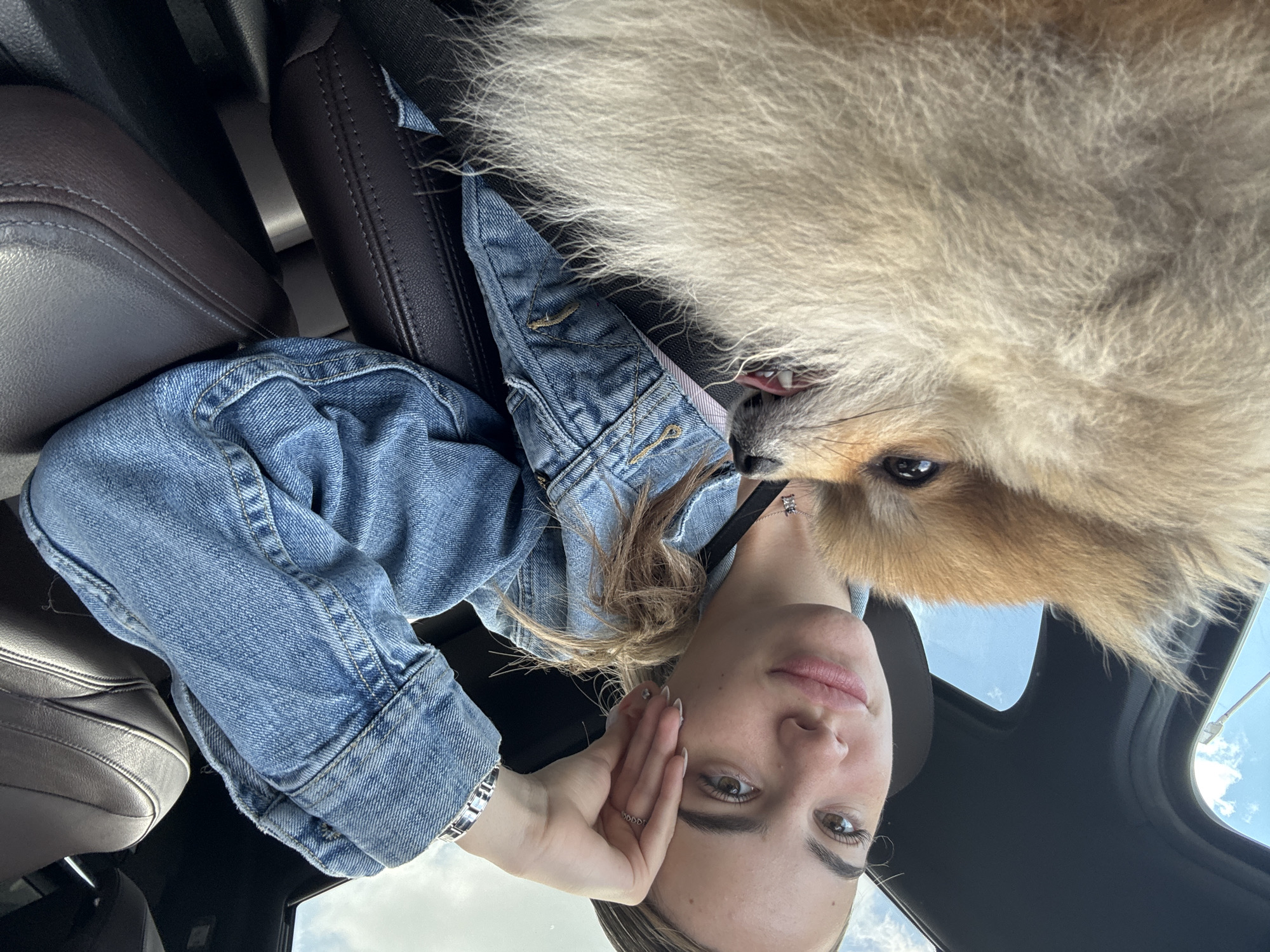 An originally rotated image, when viewed upright, shows a young woman and a fluffy light brown dog (likely a Pomeranian) in a close-up selfie taken inside a car. The woman, with light brown hair and wearing a denim jacket, looks directly at the camera with a calm expression, her hand gently touching her cheek. A silver-toned necklace is visible around her neck. The dog, with its abundant fur, is positioned very close to the woman's face, possibly resting on her shoulder or lap. One of its eyes is visible, looking towards the camera, and its mouth is slightly open, showing a hint of teeth and tongue.
The setting is a car interior, with dark brown leather seats visible to the side and below the subjects. Above them, a sunroof or window reveals a bright, cloudy blue sky, indicating it is daytime. The overall atmosphere appears relaxed and content. There is no visible text in the media file. The scene is located in Sofia, Bulgaria.