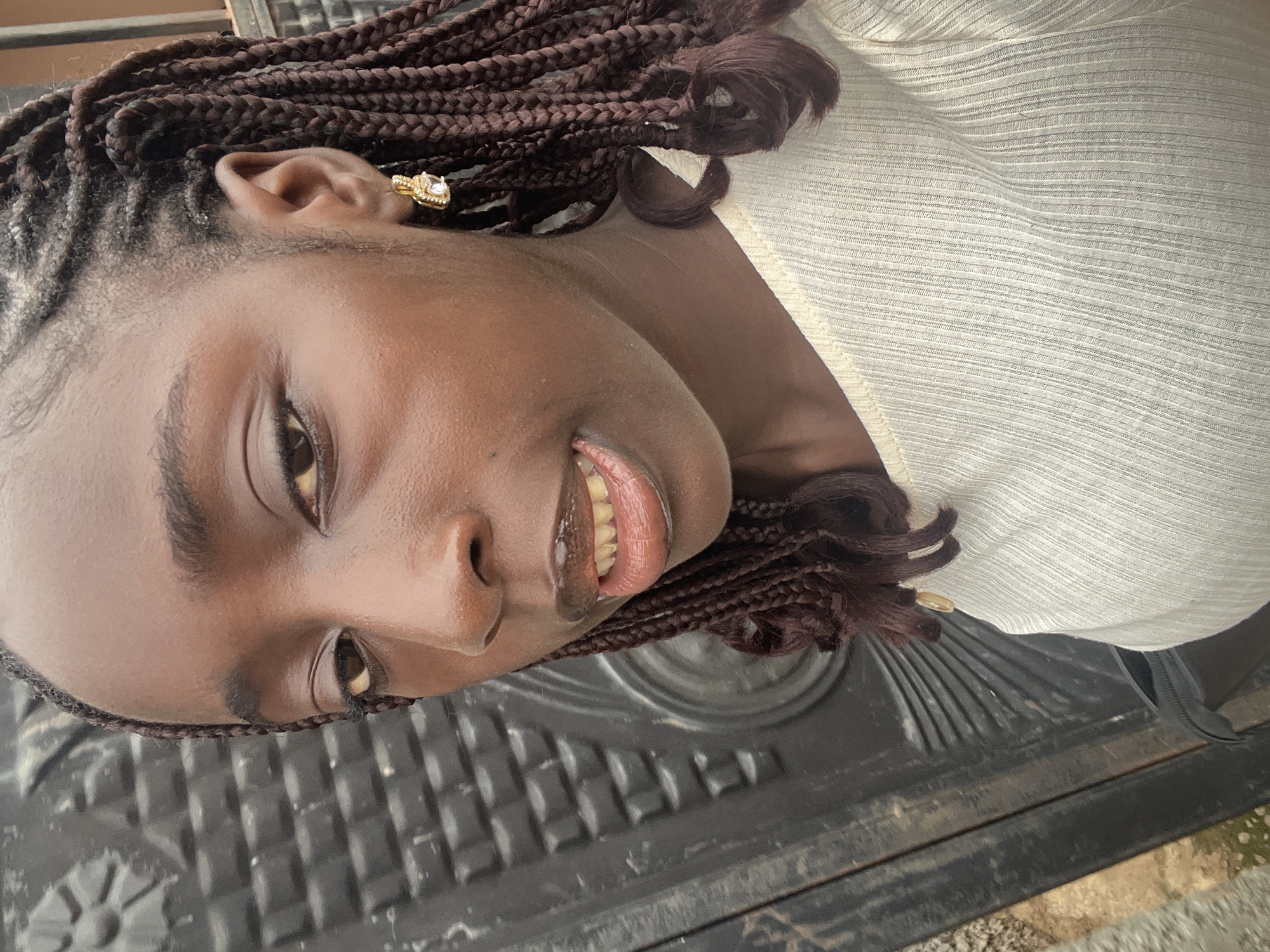 A close-up portrait features a young woman with radiant dark skin, captured with a joyful expression. Her hair is styled in neat, dark brown box braids that frame her face and extend over her shoulders. She wears a striking gold-toned earring, adorned with a prominent square, clear gemstone, in her right ear. Her smile is wide and genuine, revealing her upper teeth, and her eyes are bright as she looks slightly upward and to her side. She is dressed in a light, possibly cream or off-white, top with a subtle horizontally ribbed texture. The background is a dark, heavily textured surface, resembling a patterned metal or durable plastic panel, possibly an exterior door or gate. The natural lighting on her face is bright, highlighting her features. This image was captured in Abuja, Nigeria.