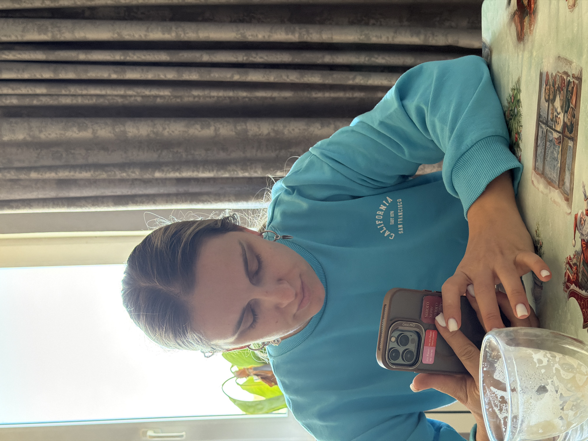 A young woman with fair skin and shoulder-length brown hair is seated at a table, looking down at a smartphone in her hands. She is wearing a light blue sweatshirt with "CALIFORNIA EAST 1976 SAN FRANCISCO" printed on it. The phone has a dark brown case with stickers that include Russian text and the phrase "CASE PRO". One of the stickers reads "AhAl это будет не легко но это будет того стоить Много Хочу МНОГО". Her fingernails are painted white. There is a half-empty glass with a foamy liquid near her phone on the table. The table is covered with a patterned tablecloth, and behind her is a window with natural light and a blurred green plant on the windowsill. In the background are gray curtains. The location is Chaikovsky, Russia.
