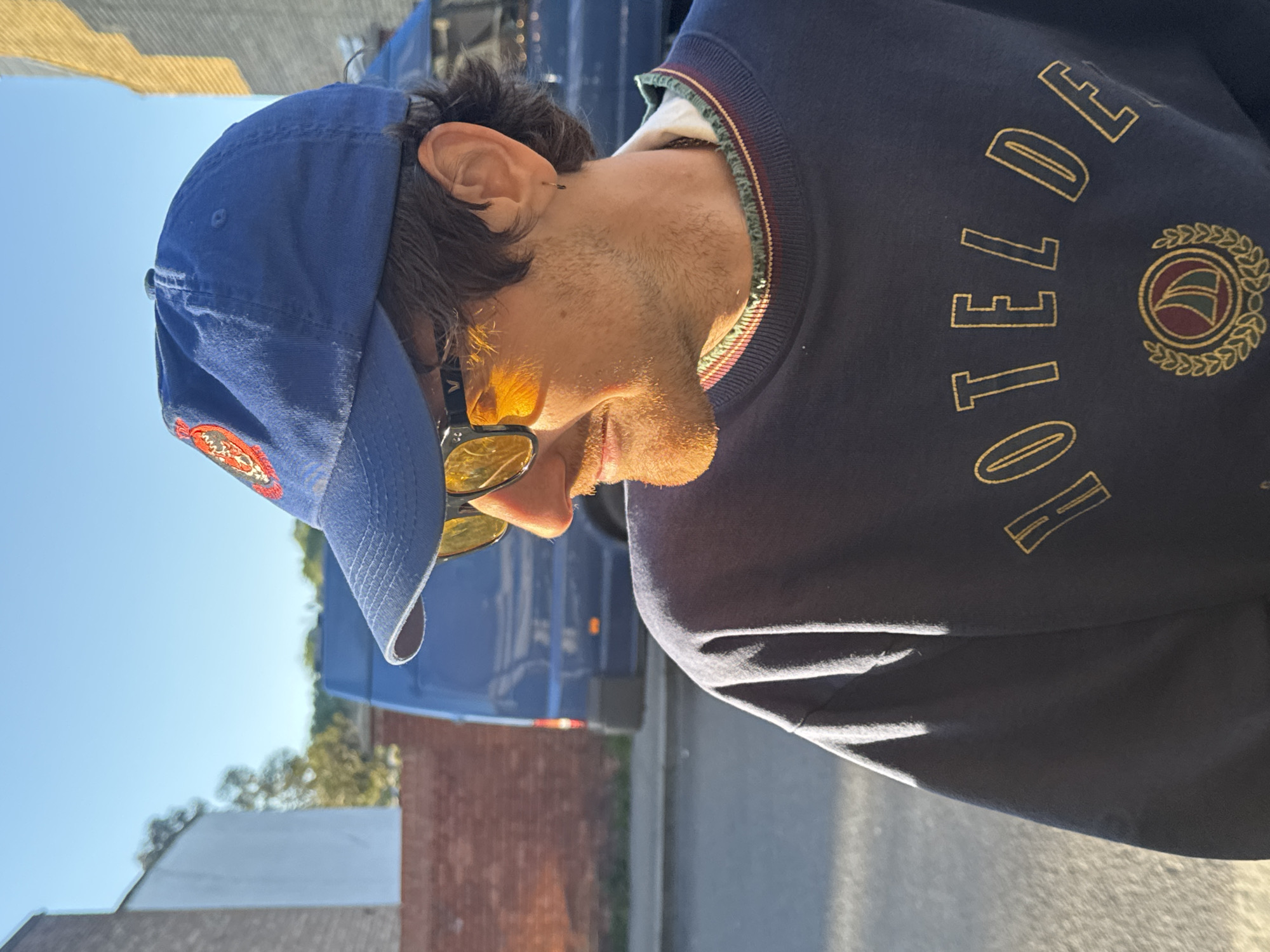The image displays a man in profile, oriented sideways (rotated 90 degrees clockwise). He is outdoors under bright sunlight, likely during the day, given the clear blue sky and strong shadows. The man has short dark hair, stubble on his face, and is wearing a blue baseball cap with an indistinct red and orange logo. He also wears yellow-tinted sunglasses and a small silver earring in his right ear.
He is dressed in a dark blue crew-neck sweater that features yellow embroidery. The embroidery includes the partial text "HOTEL DE" and a circular crest design. This crest consists of a laurel wreath encircling an emblem, possibly a sailing ship or flag. The sunlight casts a warm, golden glow on the right side of his face and sweater.
In the background, a large blue commercial vehicle, possibly a van or truck, is visible. Further back, there are distant buildings, one with a reddish-brown brick facade, and green foliage, suggesting an urban or suburban street setting. No specific activity or emotion is clearly discernible from the profile view. The scene is consistent with a street view in a city like London, United Kingdom.