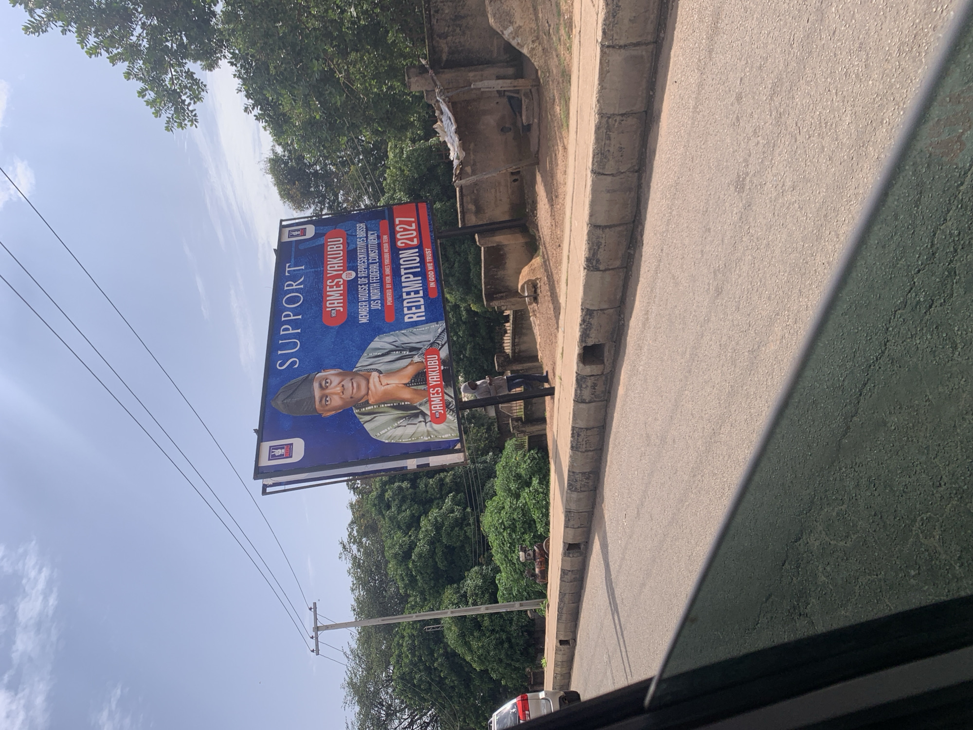 The image shows a billboard featuring James Yakubu, a candidate for the House of Representatives for the Bassa-Jos North Federal Constituency. The billboard has the text "SUPPORT JAMES YAKUBU" prominently displayed, along with "REDEMPTION 2027" and "IN GOD WE TRUST". Yakubu is shown in traditional attire wearing a cap. The billboard is situated outdoors, beside a paved road with a concrete sidewalk. Green trees and a cloudy sky are visible in the background. There are power lines and a utility pole in the foreground. The overall scene appears to be a street in Jos, Nigeria, possibly during the daytime given the lighting.