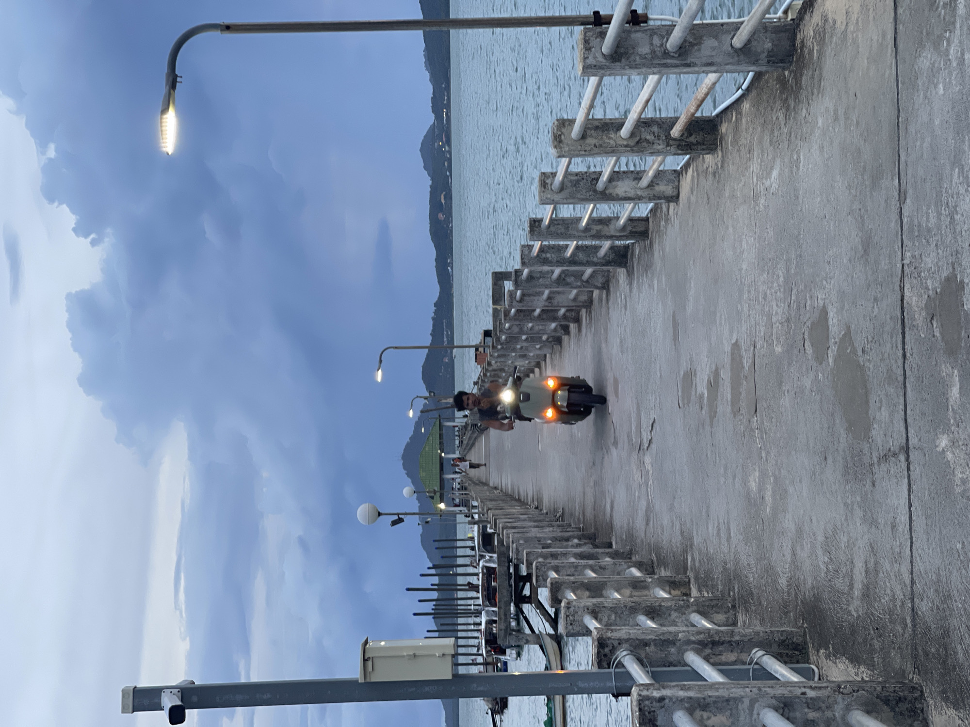The image depicts a person riding a scooter down a pier. The scene appears to be a waterfront location, likely a harbor or a coastal area, given the presence of the sea and distant hills. The time of day is likely dusk or early evening, as indicated by the soft lighting from streetlights on the pier and the somewhat overcast sky. The weather appears to be cloudy. The pier itself is made of concrete and has a railing with metal bars. There are several streetlights along the pier, illuminating the path. In the background, hills covered in vegetation can be seen rising from the water, and there are some buildings visible along the coastline. The scooter has its headlights on, casting a glow on the wet-looking pavement. The person on the scooter is looking forward and appears to be alone. The overall atmosphere is serene and tranquil. No visible text is present in the image. The location context of Pa Khlok, Thailand, suggests a tropical coastal environment.