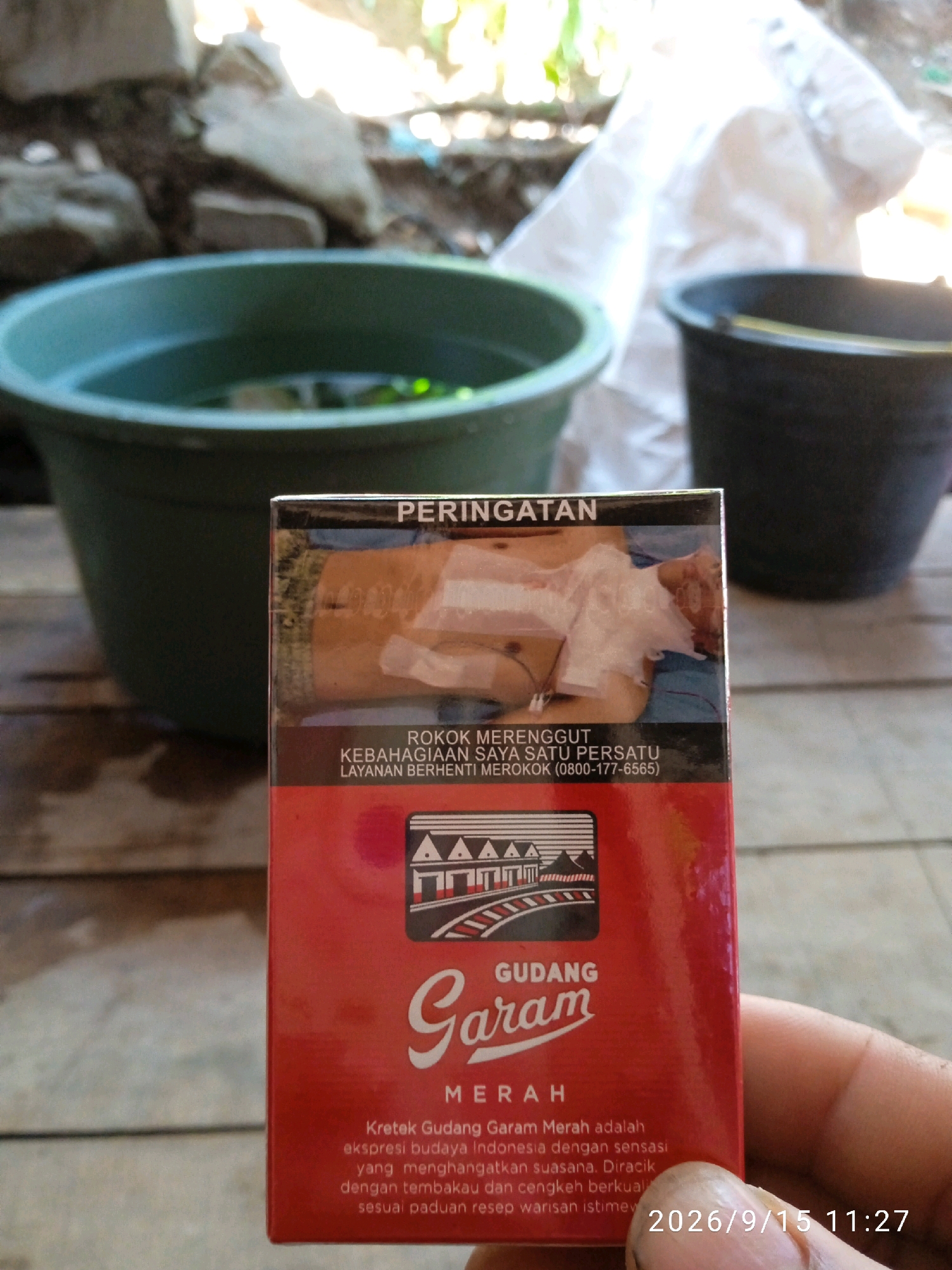 A close-up shot features a person's thumb and index finger holding a red cigarette pack, partially obscuring the bottom right corner. The cigarette pack has a warning label on the top half depicting a graphic of a person bandaged, with text above and below. The brand name "GUDANG GARAM MERAH" is prominently displayed on the lower half of the pack. The background is blurred, showing an outdoor setting with green and black buckets and what appears to be natural light filtering through. The timestamp "2026/9/15 11:27" is superimposed on the bottom right of the image. The scene is set in Kelurahan, Indonesia.