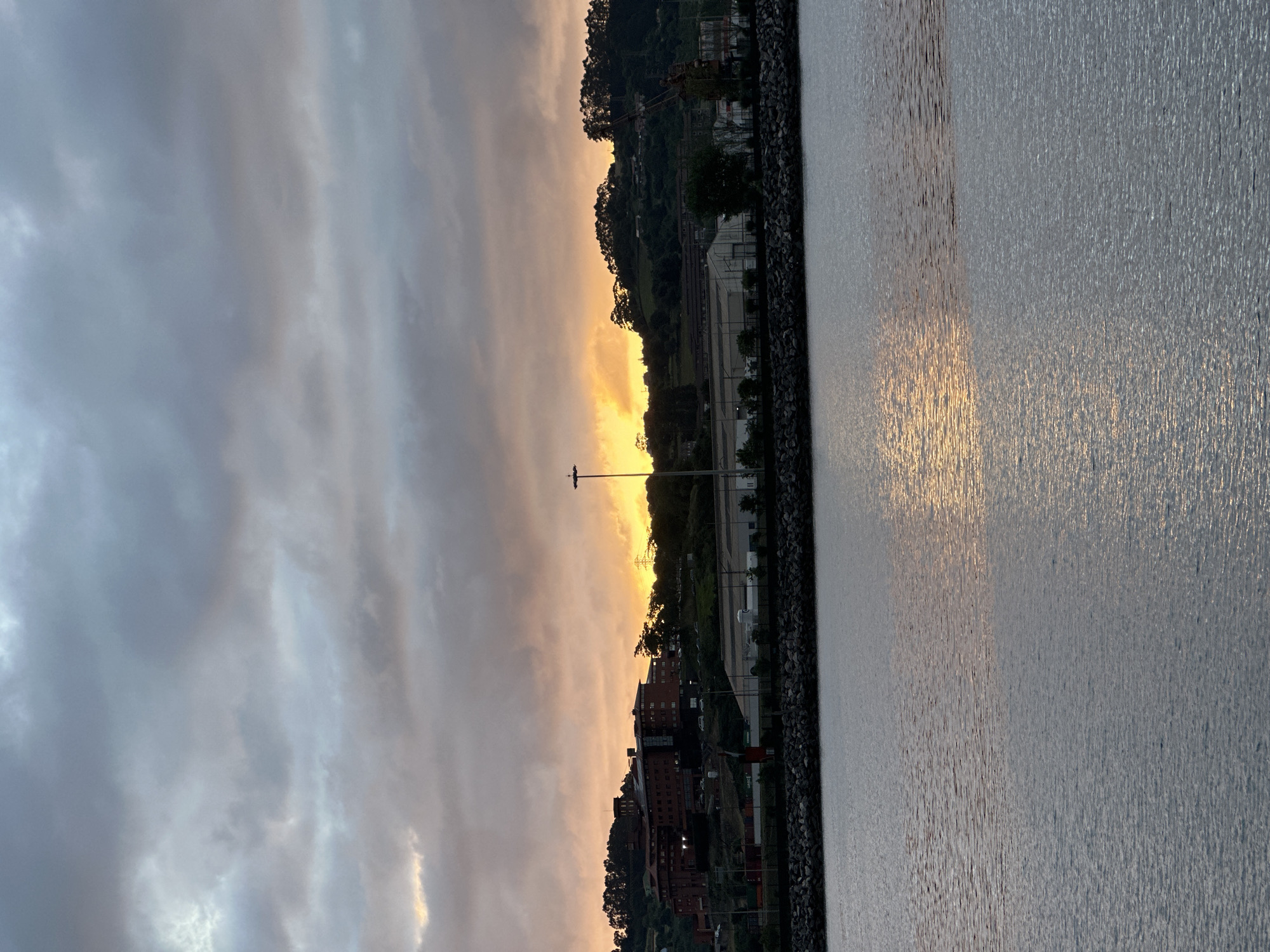 The image depicts a waterfront scene at dusk or dawn, likely in the city of Gijón, Spain, with the sun setting or rising behind a hill covered in trees and buildings. In the foreground, a calm body of water reflects the muted colors of the sky, which is dominated by gray and orange clouds. Along the horizon, a stone embankment separates the water from the land. Beyond the embankment, a large, multi-story brick building is visible on the side of the hill, with other industrial or commercial structures closer to the water. A tall light pole stands prominently on the right side of the image, and a crane can be seen further to the right, suggesting an industrial or port area. The overall atmosphere is tranquil and slightly overcast, with the warm hues of the sky providing a touch of color against the muted tones of the landscape. There are no people visible in the image.