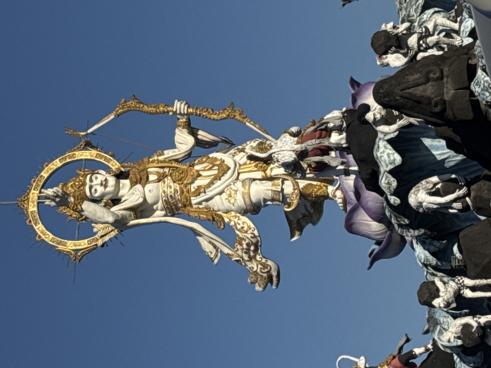 The image shows a statue of Arjuna with bow and arrow on a float during the Denpasar Festival in Bali, Indonesia. The statue is light-colored with gold trim and stands out against a clear blue sky. The float is decorated with dark-colored figures.