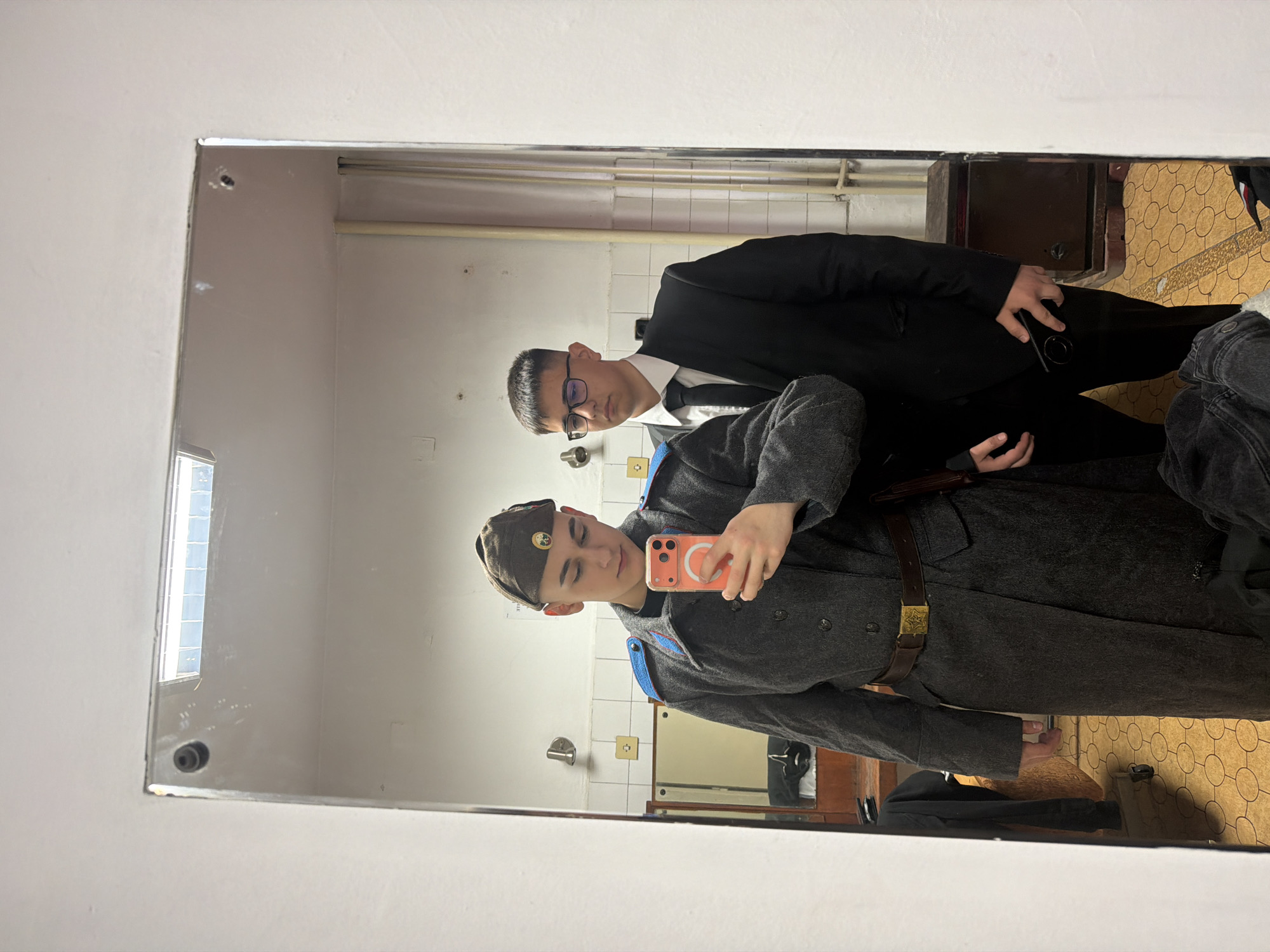 Two men are reflected in a mirror. The man in the foreground wears a military-style gray overcoat with a brown leather belt and a side cap with a badge. He holds a phone in his hand, taking a selfie. The man behind him wears a black suit with a white shirt and tie. The room appears to be a dressing room or restroom, with white tile walls and visible pipes. The floor is covered in patterned linoleum. A cabinet is visible in the background. The location is consistent with the style of architecture and design often found in older buildings in Panagyurishte, Bulgaria.
