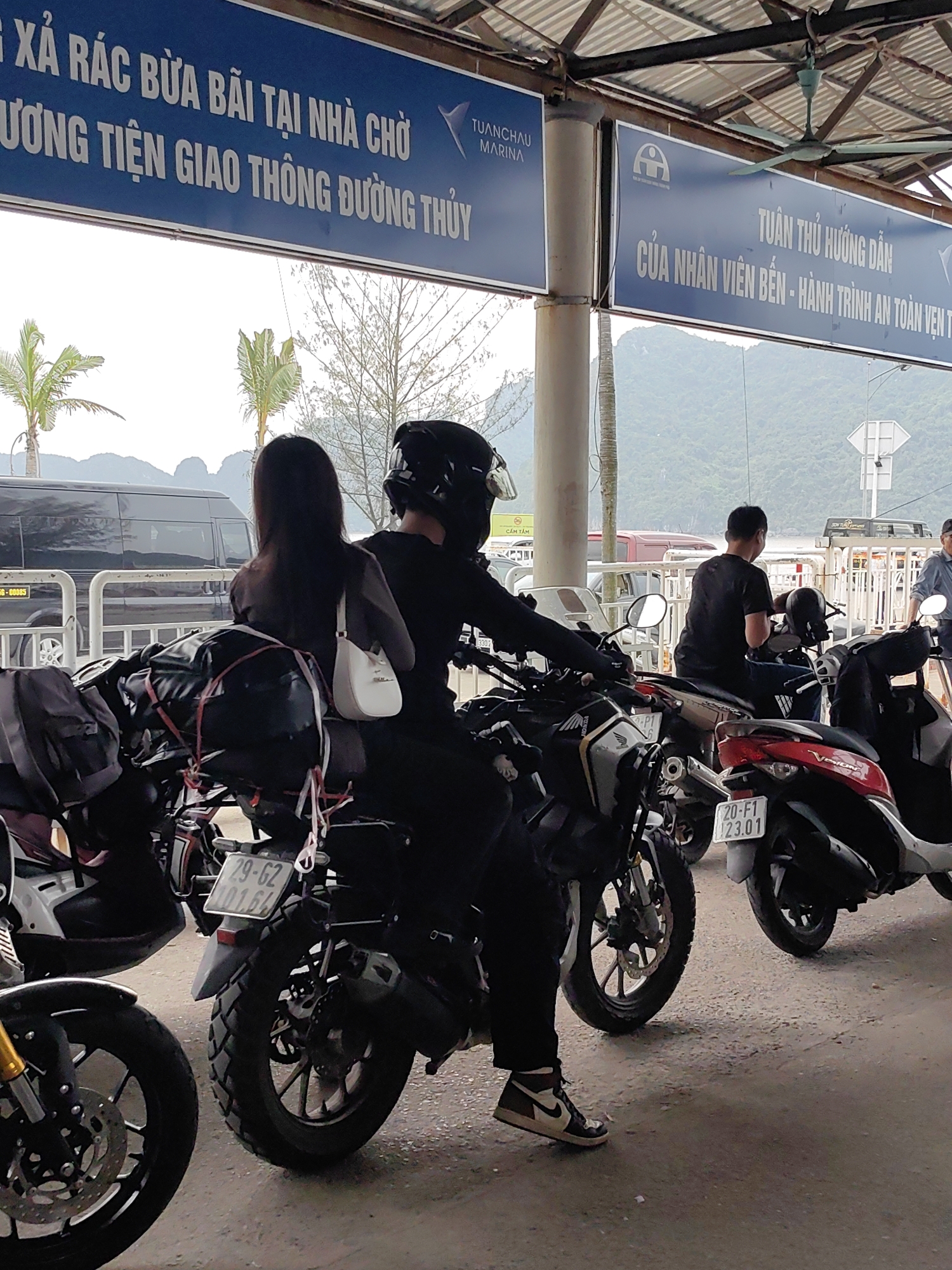 The image shows a scene at the Tuan Chau Marina in the city of Hải Phòng, Vietnam. Several motorcycles are parked under a covered area with signage in Vietnamese. One sign reads "TUANCHAU MARINA" and includes information about proper disposal of trash and safety instructions. A couple is on one of the motorcycles, with the rider wearing a helmet and the passenger carrying a white purse. The motorcycle is loaded with luggage. Other people are visible nearby. The background features mountains and a cloudy sky.