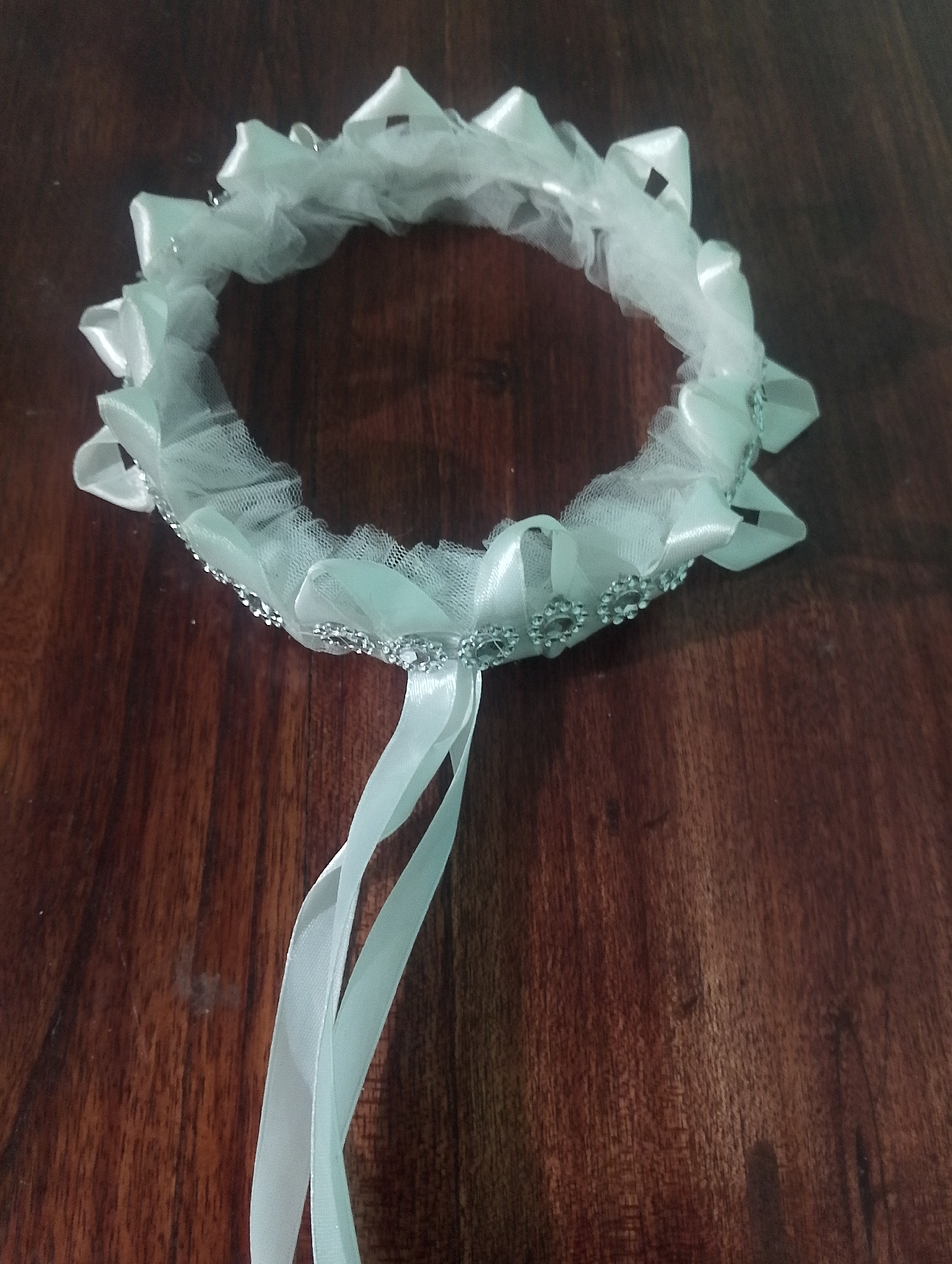 A white garter with ribbons and rhinestones is lying on a dark wooden surface. The garter is a circular band of white tulle and satin ribbons, embellished with small, sparkling rhinestones along the bottom edge. Two white ribbons hang down from the garter. The background is a dark, grainy wooden surface, possibly a table. The image is taken from a slightly elevated angle, looking down at the garter. The lighting is soft, casting a slight shadow from the garter onto the wood. There are no people or any other objects visible in the frame.