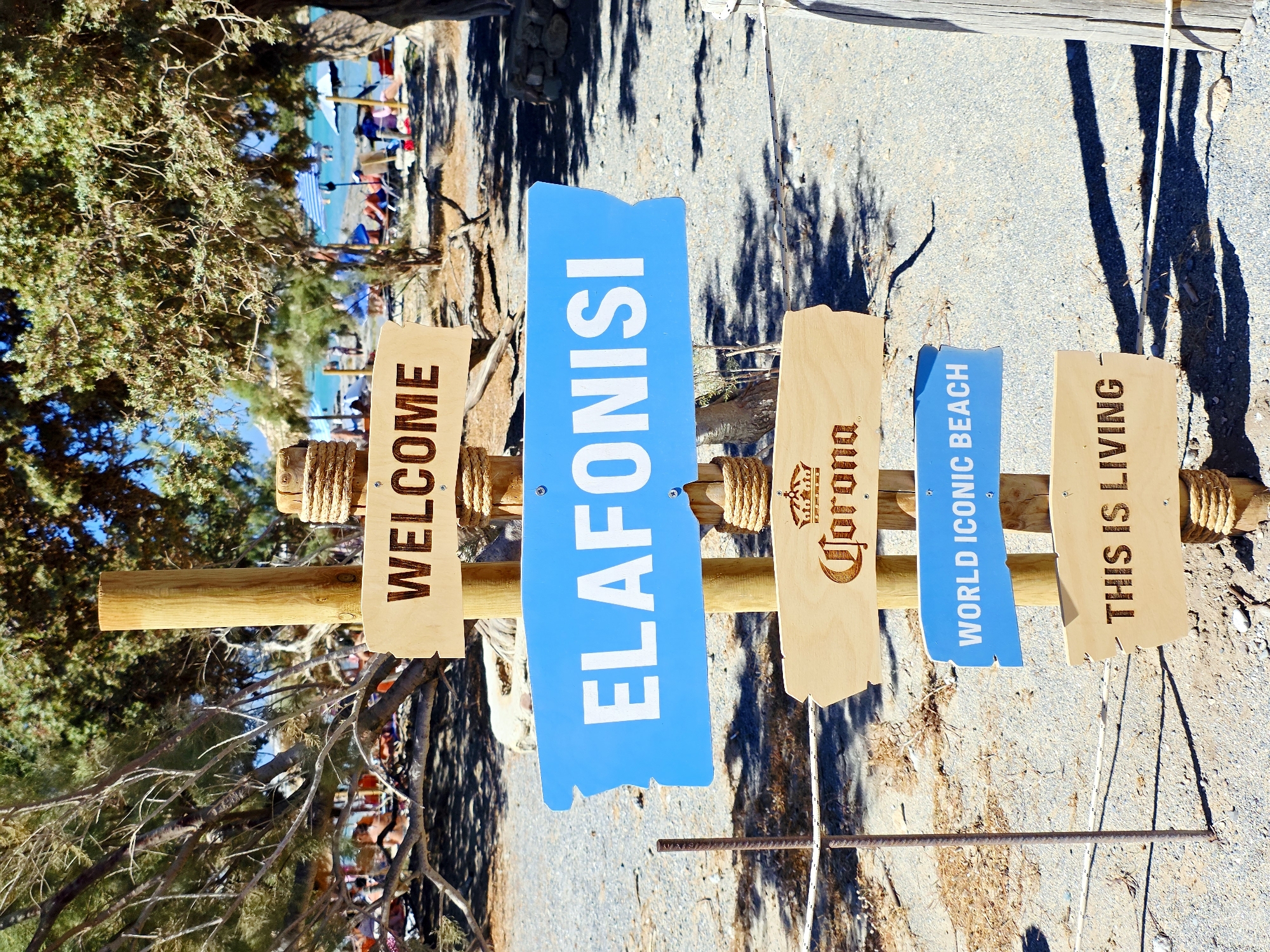 A wooden post with multiple signs stands on a sandy beach in Greece. The signs are directional and informational. The top sign is made of light-colored wood with dark lettering that reads "WELCOME". Below this, a bright blue sign with white lettering points to the right and reads "ELAFONISI". The next sign down is also light-colored wood with dark engraving of a crown and the word "Corona". A smaller blue sign with white lettering reads "WORLD ICONIC BEACH". The bottom sign is light-colored wood with dark lettering that reads "THIS IS LIVING". The signs are attached to the wooden post with screws. The beach is populated with people relaxing under umbrellas, and the sea is visible in the background, appearing turquoise. Trees with green foliage provide shade. The ground is covered in gravel and sand.
