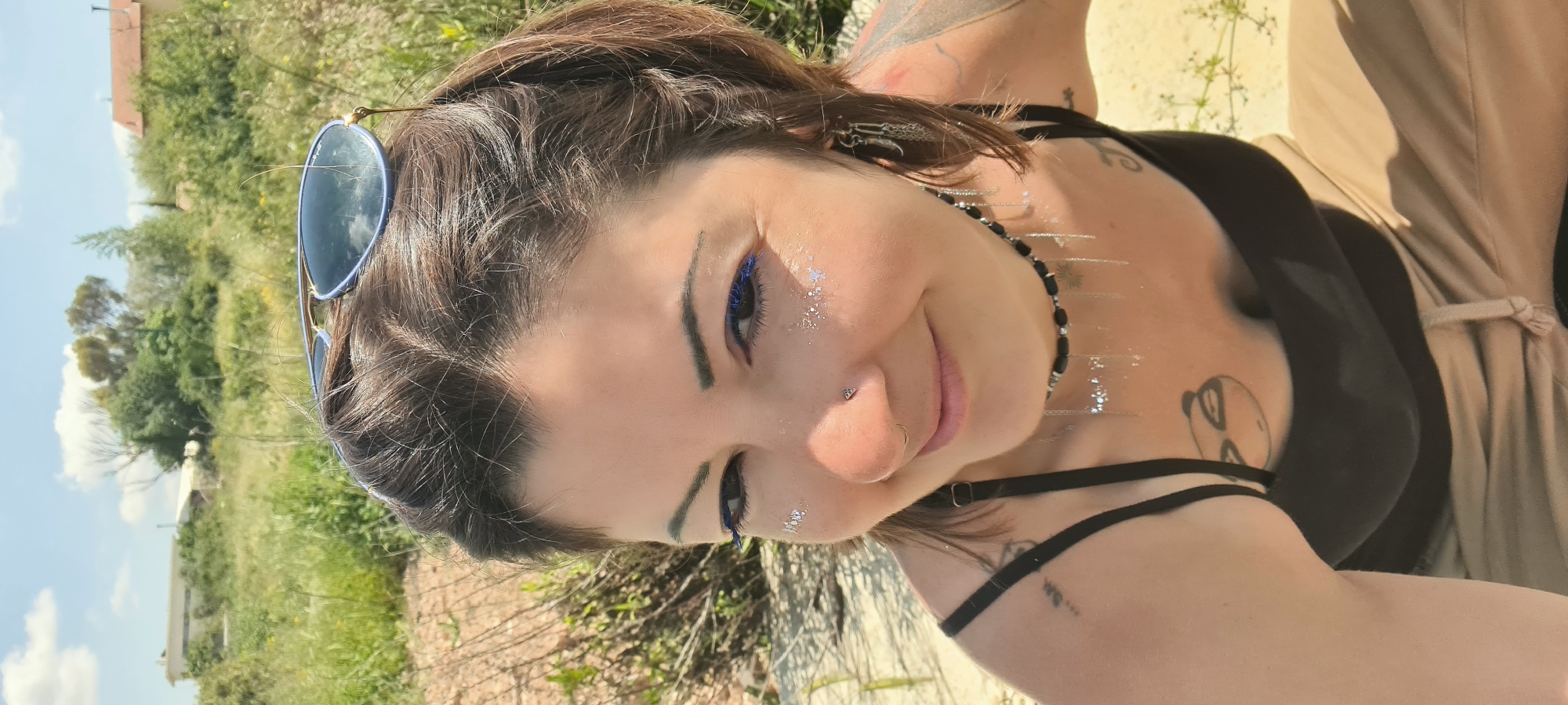 A medium shot of a smiling woman, positioned at an angle, with a landscape in the background. The woman has dark hair, fair skin, and visible tattoos on her upper chest and arms. She is wearing dark eye makeup and has glitter on her face and chest. A pair of sunglasses rests on top of her head. She is wearing a black tank top and what appears to be a beige coat or robe, fastened with a tie. She is also wearing a beaded necklace.
The background includes green vegetation and what appears to be a building. The location is in the city of Lachish Regional Council, Israel.