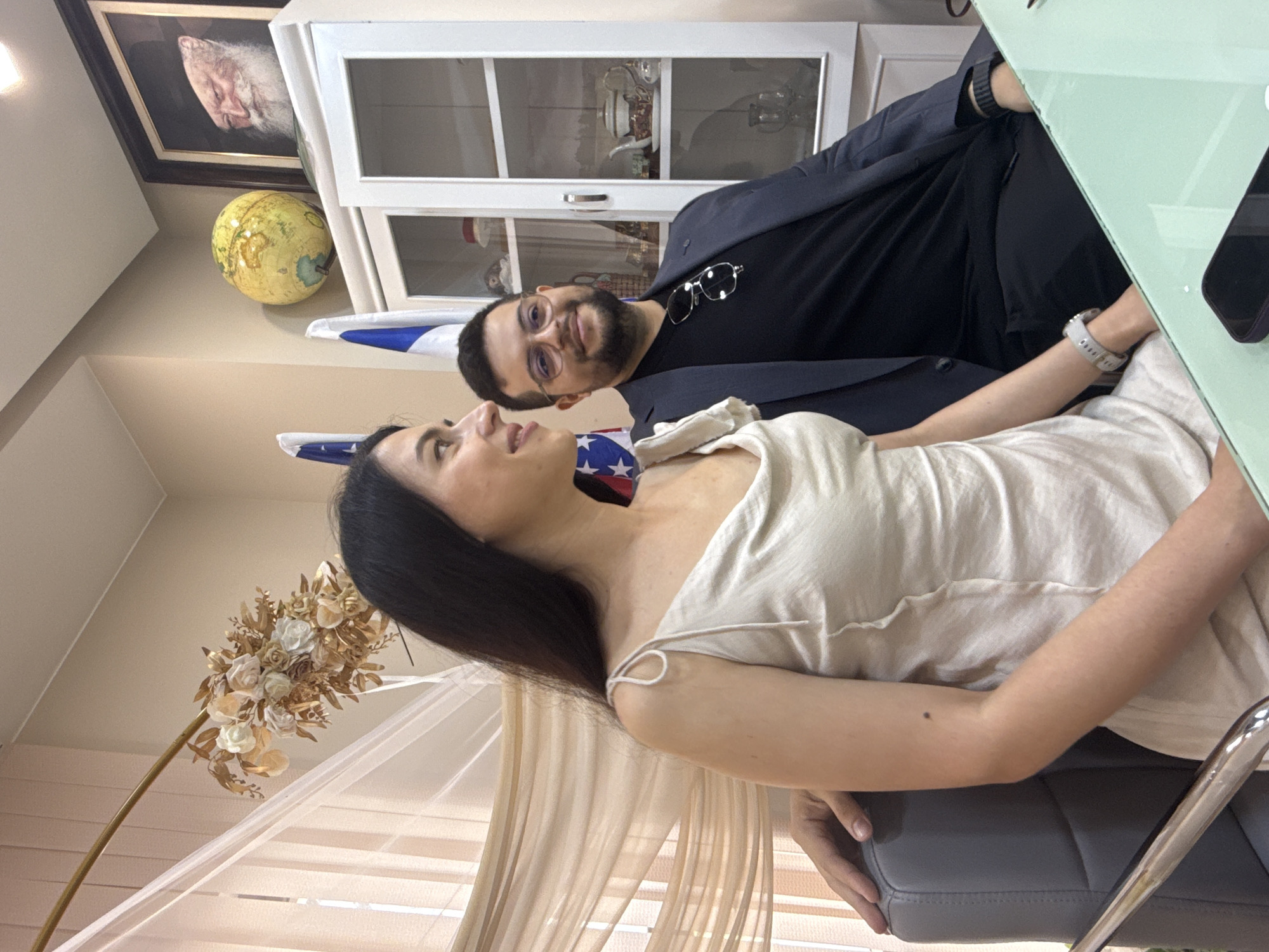 An indoor photograph, rotated 90 degrees clockwise, captures a man and a woman in what appears to be an office or decorated room in Rishon LeZion, Israel.
The man, with a beard, short dark hair, and eyeglasses, is positioned slightly behind the woman and looks directly at the camera with a gentle smile. He wears a black t-shirt underneath a dark blazer. A pair of silver-rimmed sunglasses is tucked into his t-shirt collar.
To his right, a woman with long dark hair is seated, looking towards the left side of the frame, not at the camera. She wears a light-colored, possibly off-white or beige, sleeveless top or dress. Her right arm rests on a light green table, and she wears a light grey smartwatch. Her left hand is visible, resting on the armrest of a dark grey office-style chair.
The background behind the man features a white cabinet with glass doors displaying decorative items, and above it, a framed portrait of an older man with a white beard. To the left of the cabinet, a vintage-style globe is visible. An Israeli flag is draped behind the man. Behind the woman, a sheer, light-colored curtain or fabric drape is visible. To her left, a decorative floral arrangement with white and gold flowers and foliage stands on a golden arched frame. A partial American flag is also visible behind the woman's neck area.
The scene suggests a posed photograph, possibly during an interview or meeting, given the office-like elements combined with national flags. The lighting is bright, indicating a well-lit indoor environment. There is no visible text in the image.