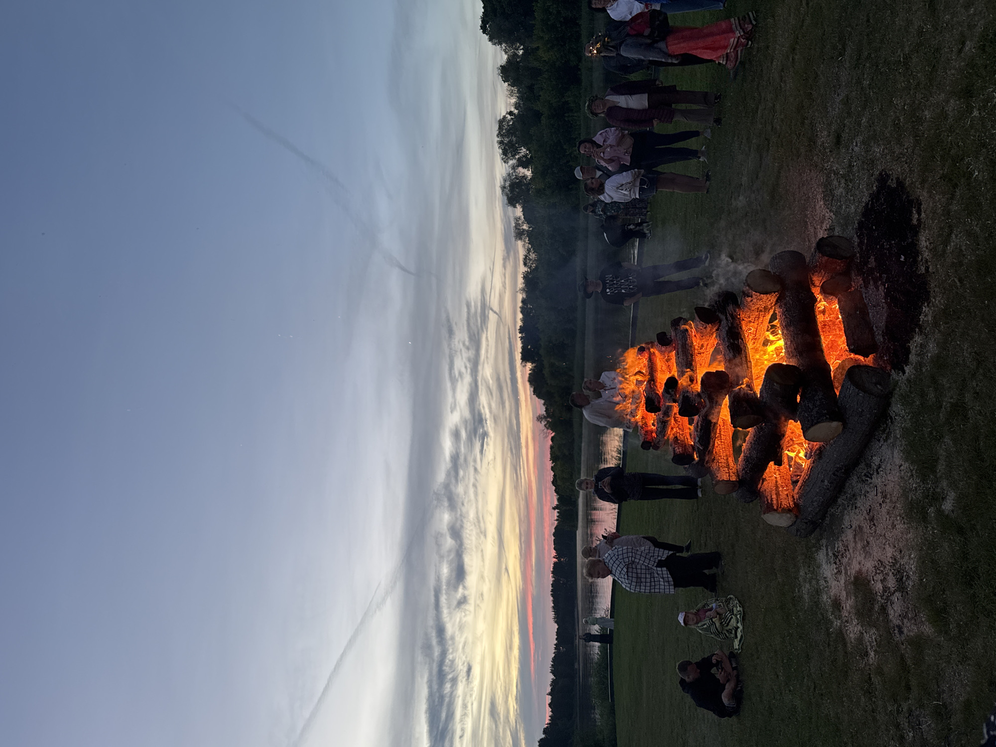 The image depicts an outdoor scene at dusk, featuring a large bonfire constructed from logs. Several people are gathered around the bonfire and in the surrounding grassy area. The bonfire is actively burning, casting a warm glow on the surrounding logs and the faces of some onlookers. In the background, a body of water, likely a river, is visible, bordered by a treeline. The sky above displays a twilight gradient with some cloud formations. The overall atmosphere suggests a communal gathering, possibly a celebration or event, taking place in Carnikava, Latvia, given the context of the location.