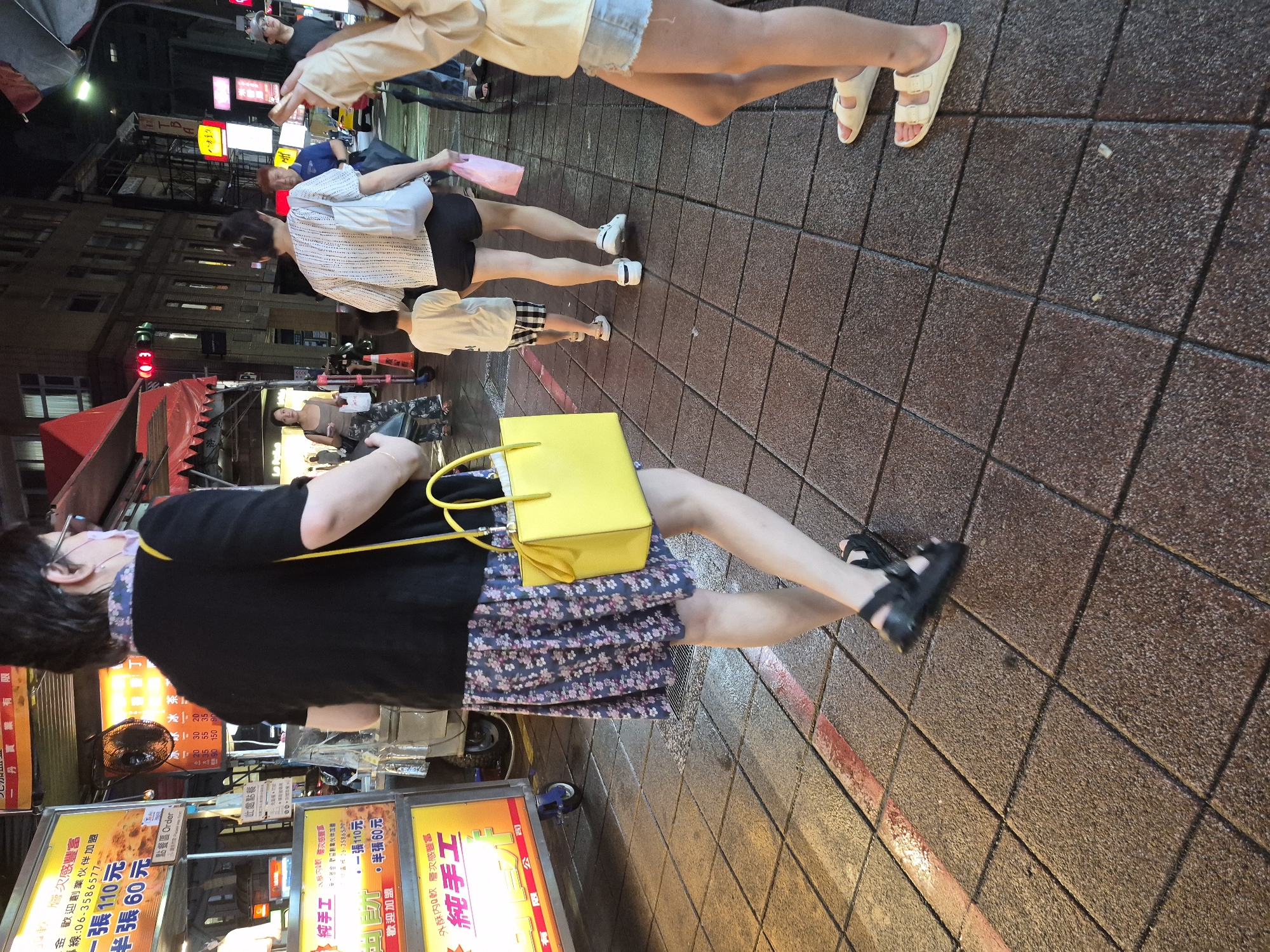 The image captures a street scene in Taipei, Taiwan, at night. The foreground is dominated by a pedestrian walkway paved with textured brown square tiles, some of which appear wet. A woman, viewed from behind, is walking away from the camera. She has short dark hair, wears glasses, and is dressed in a black top and a floral patterned skirt. She carries a bright yellow handbag on her right shoulder. Her feet are shod in black platform sandals. To her left, a food cart is visible, displaying signs with Chinese characters and prices, such as "半張 60元" (half portion 60 NT dollars) and "張 110元" (whole portion 110 NT dollars). The cart has a yellow sign with "純手工" (handmade) prominently displayed. Behind the woman in the foreground, other pedestrians are visible. A child in a white t-shirt and plaid shorts walks ahead of a woman in a white patterned shirt and black shorts. Further in the background, more people and buildings suggest an urban environment, with some streetlights and neon signs illuminating the night.