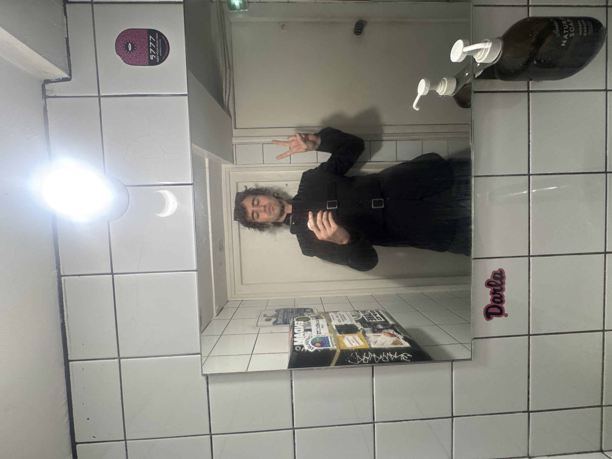 Here is a detailed description of the image:
A man with curly dark hair is taking a selfie in a mirror in a tiled bathroom or restroom. He is wearing a black outfit with straps across the torso and making a peace sign with his left hand. The mirror reflects the interior of the bathroom.
The bathroom is tiled in white, square tiles with dark grout. There is a light fixture above the mirror and a crescent moon drawing is visible on the tiles. There is a sticker with the text "5777 KEEP YOUR HEAD HIGH" on the top left.
The mirror is affixed to the wall and has multiple stickers on its surface. Two soap bottles are attached to the tile wall above the mirror. On the right side of the tile wall next to the mirror is a pink sticker with the name "Darla".
Given the graffiti and the type of stickers, the location could potentially be a public bathroom or a restroom in an alternative space in Paris.