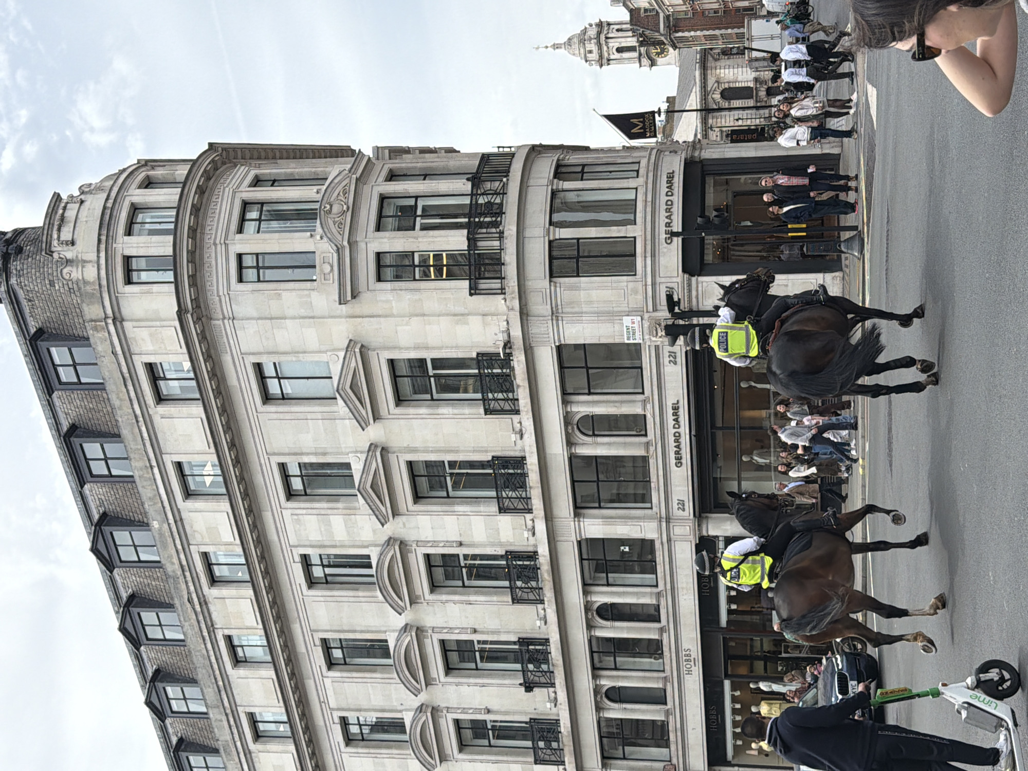 This image shows a street scene in London, United Kingdom. The building visible in the background has the signage "GERARD DAREL" and the address "221." There are also signs that say "HOBBS" and "MADDOX GALLERY" visible. A "REGENT STREET W1" street sign is also present. In the foreground are two mounted police officers on horseback and other people. A scooter and a person standing nearby are on the right-hand side of the frame. Another person is holding up a phone to take a picture in the upper right-hand corner of the frame.