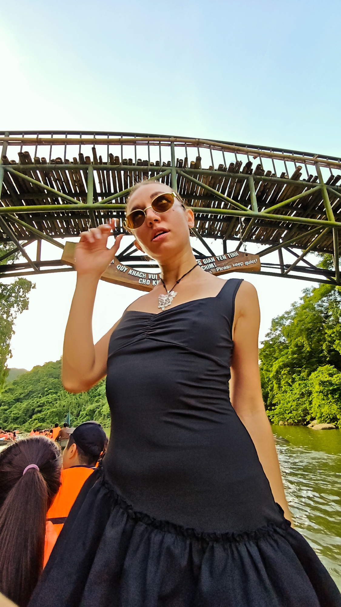 A low-angle, wide-shot portrait shows a woman in a black dress standing on a boat in a body of water. The woman is wearing sunglasses and a silver necklace with a butterfly pendant. She is looking up and to the left, with her right hand near her face. Behind her, a bamboo structure with railings and a sign is visible. The sign has Vietnamese text and the English words "Please keep quiet!". To the left and right of the structure, lush green trees are visible. In the background, other boats with people wearing orange life vests are present on the water. The scene appears to be in Yên Sơn Ward, Vietnam, likely a tourist area with boat tours.