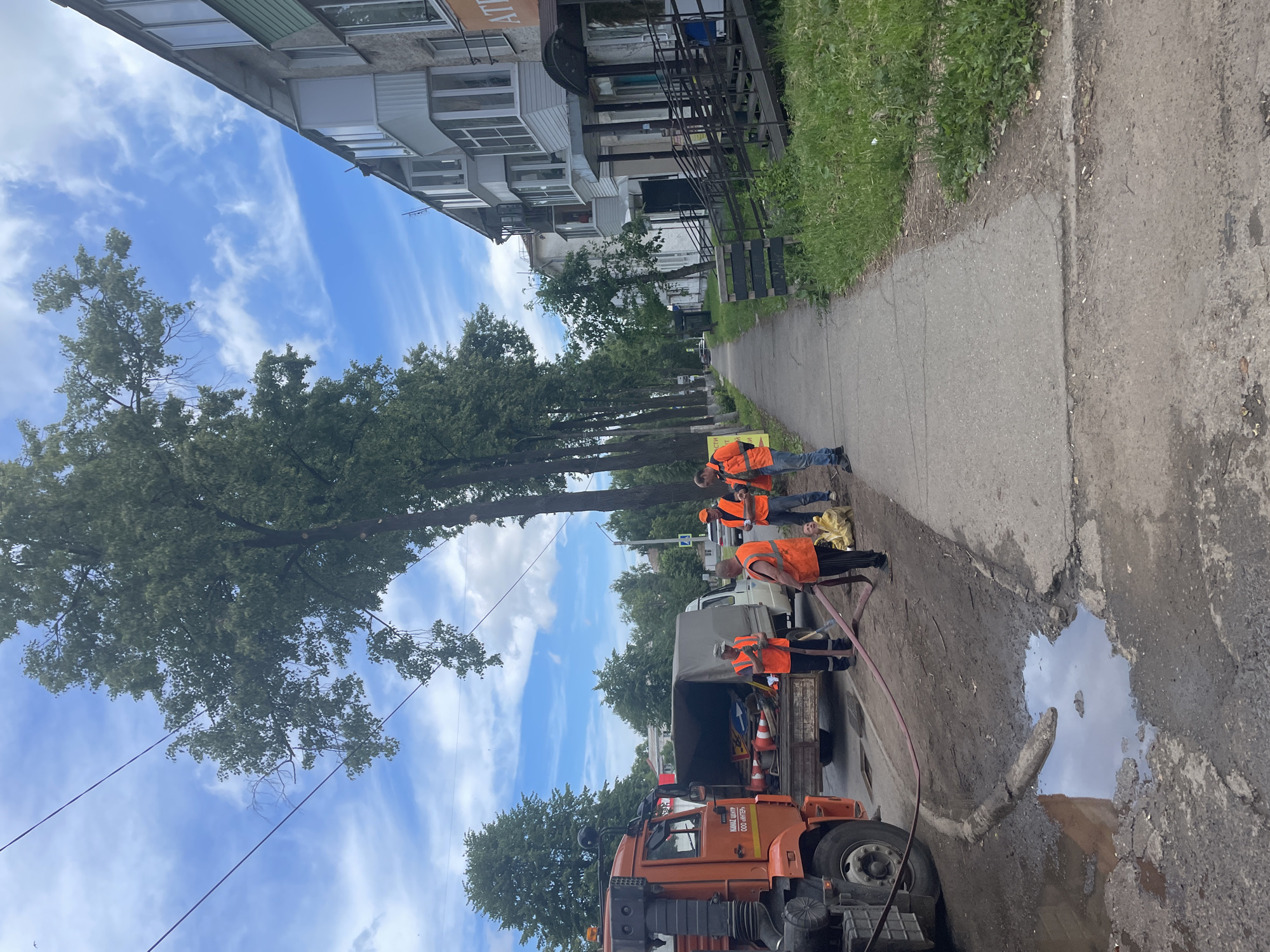 A group of utility workers in bright orange safety vests are gathered on a sidewalk in the city of Chaikovsky, Russia, near a large orange truck and a white van. They appear to be working on some sort of repair or maintenance task, as one worker is holding a large red hose connected to the truck, and another is kneeling on the ground near a muddy puddle. The scene is set on a residential street lined with trees and apartment buildings. The sky is blue with scattered clouds, suggesting a pleasant, though perhaps humid, day. The overall impression is one of an active outdoor work environment in a typical Russian urban setting.