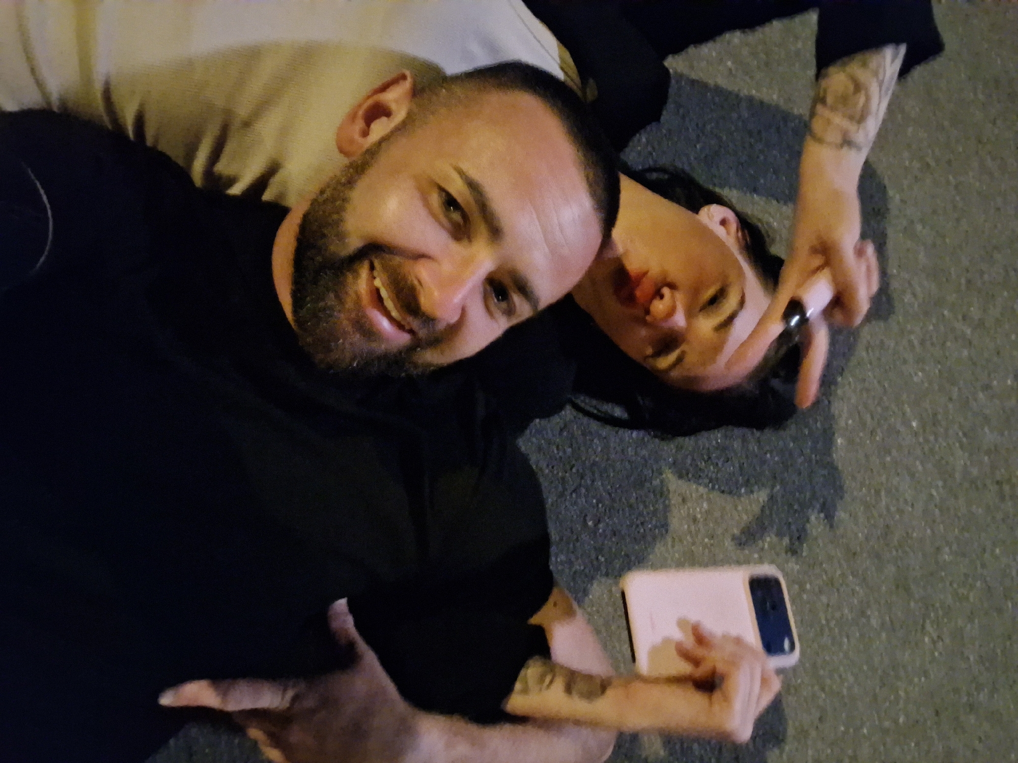 A man and a woman are lying on the ground in what appears to be a city street at night. The woman is on the left, and she is holding a pink cell phone up to take a selfie. She is making a peace sign with her left hand and puckering her lips. The man is on the right, and he is smiling at the camera. The ground is asphalt, and there are shadows on the ground, suggesting the presence of streetlights or other light sources. The woman has tattoos on her left arm. Both individuals are dressed in dark clothing.