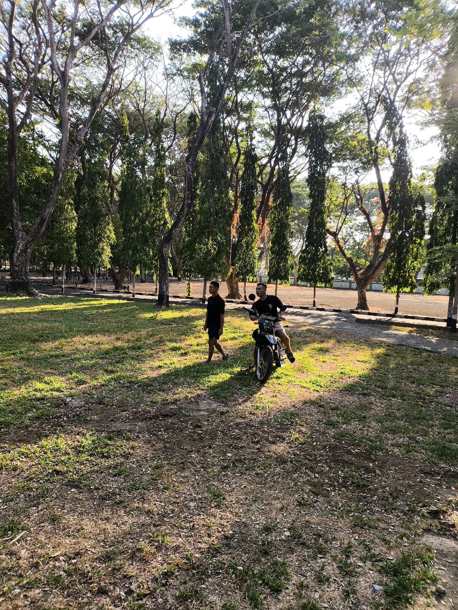 In a grassy, tree-lined area in Unknown, Indonesia, two young men are visible. One man is standing to the left, wearing a black t-shirt and shorts. The other man is seated on a motorcycle, also wearing a black t-shirt and shorts, and appears to be holding onto the handlebars. The motorcycle is parked on the grass, with a paved path behind it and a road in the background. Tall trees with dense foliage flank the scene, and the sunlight creates long shadows on the ground. The overall impression is one of an outdoor leisure activity or a casual gathering in a park-like setting.