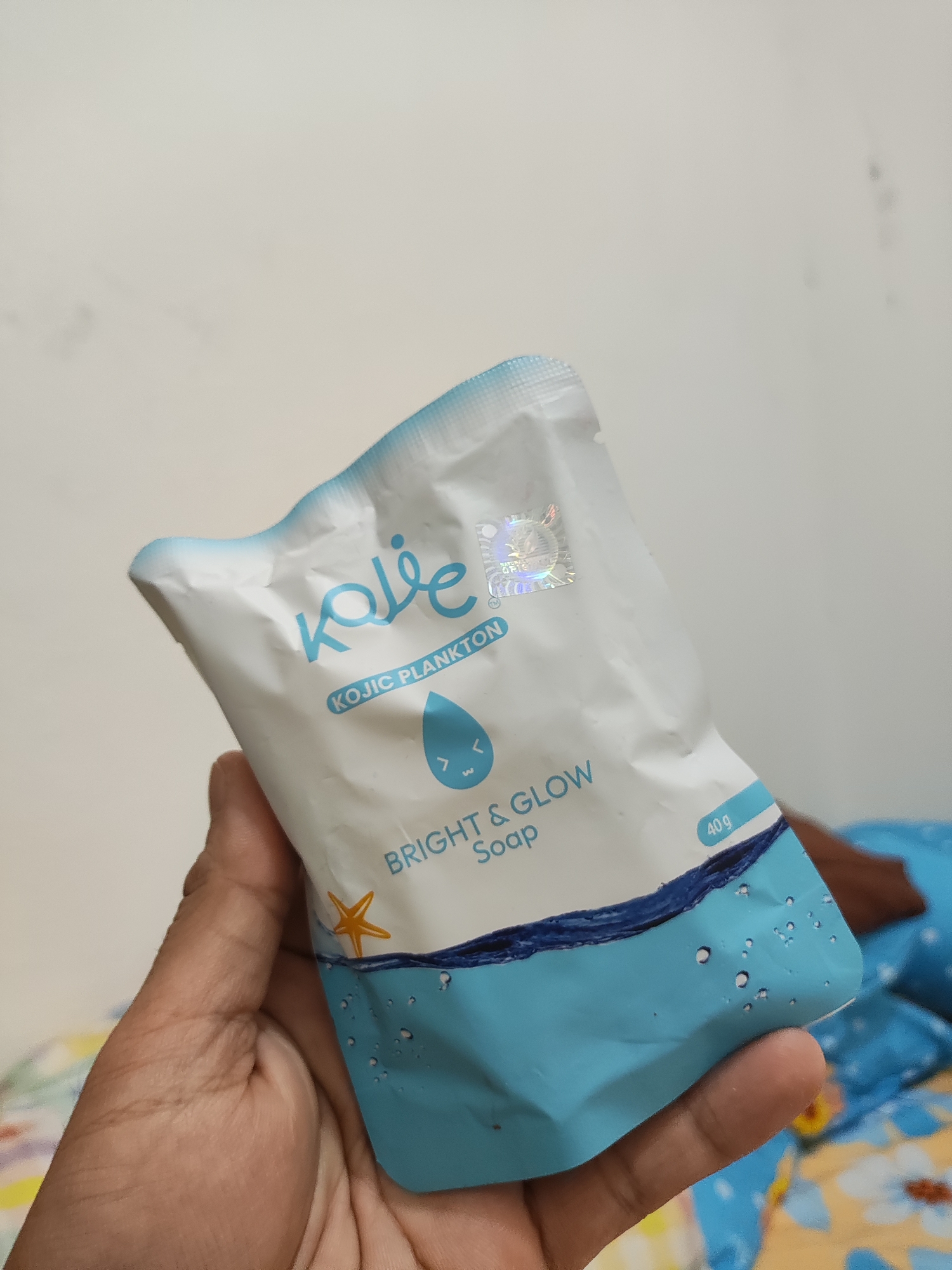 The image shows a person's hand holding a small, white and light blue packaged soap product. The packaging features blue text that reads "Kolie" and "KOJIC PLANKTON", along with a water droplet icon with a smiley face inside. Below this, it says "BRIGHT & GLOW Soap" and "40 g". A small orange starfish is visible on the lower part of the packaging. The background is a plain, lightly textured wall. This scene appears to be indoors, possibly in a residential setting in Wanajaya, Indonesia, based on the prompt.