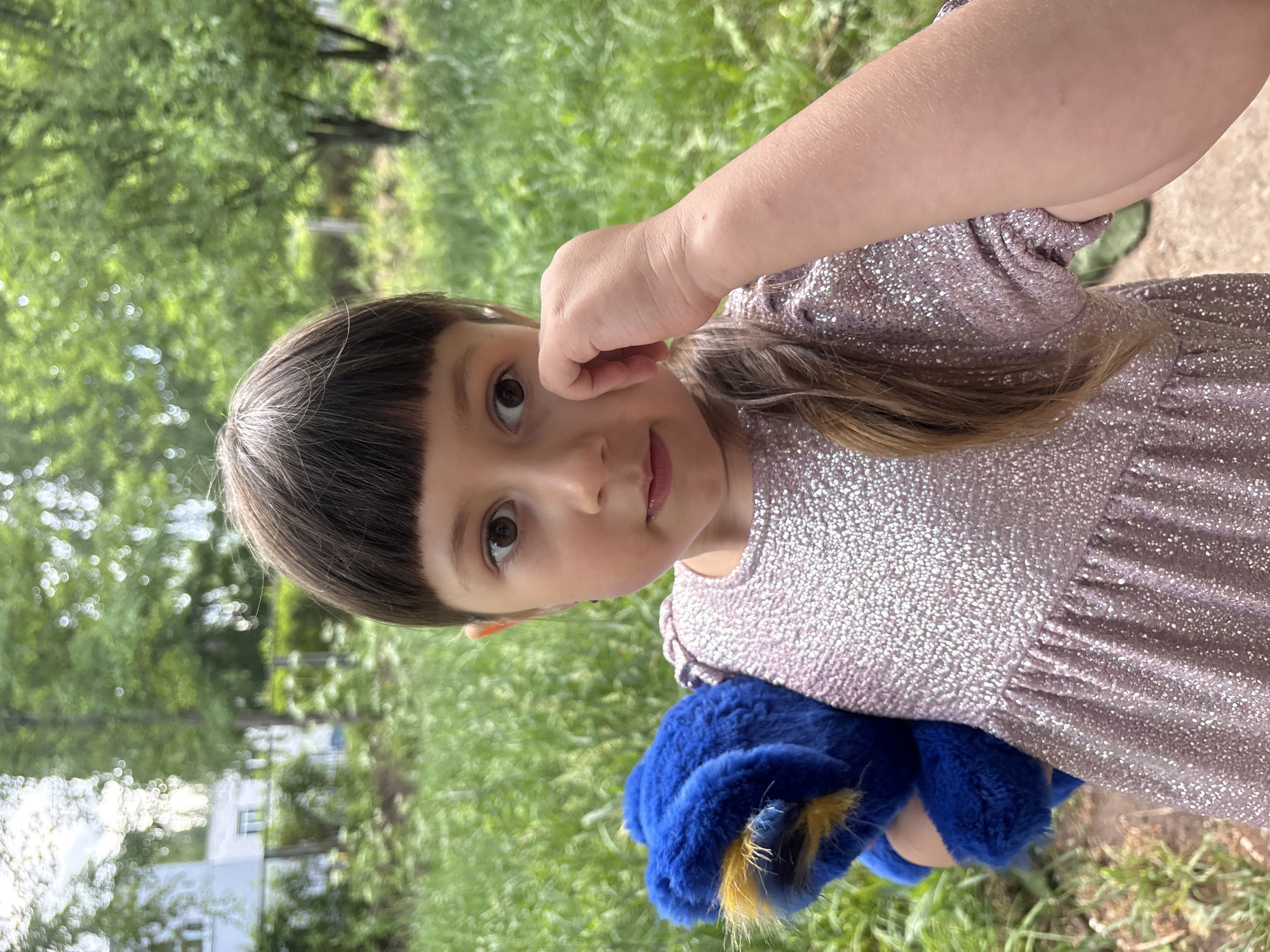 A young girl with brown hair and bangs, wearing a sparkly pink dress, stands outdoors, holding a blue stuffed animal with a yellow tail. Her right hand is raised near her face. The background includes green grass and trees, and a building can be seen in the distance. The location is Chaikovsky, Russia.