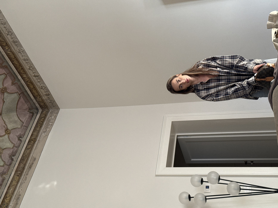 An interior photograph depicts a young adult female standing in a historic room in Venice, Italy. The female, with long brown hair featuring lighter highlights, wears a loose-fitting blue, white, and brown plaid button-down shirt. She holds a small, brown, monogrammed handbag with a leather tag in front of her, looking directly at the viewer. The room features high, unadorned white walls that meet an ornate, painted ceiling decorated with intricate patterns in pale green, pink, and gold hues, bordered by a classical frieze. To the left of the female, a modern black floor lamp with five frosted spherical light shades is positioned near a white-framed doorway, through which a grey door is visible slightly ajar. A blurred, neutral-colored furniture item occupies the lower foreground.