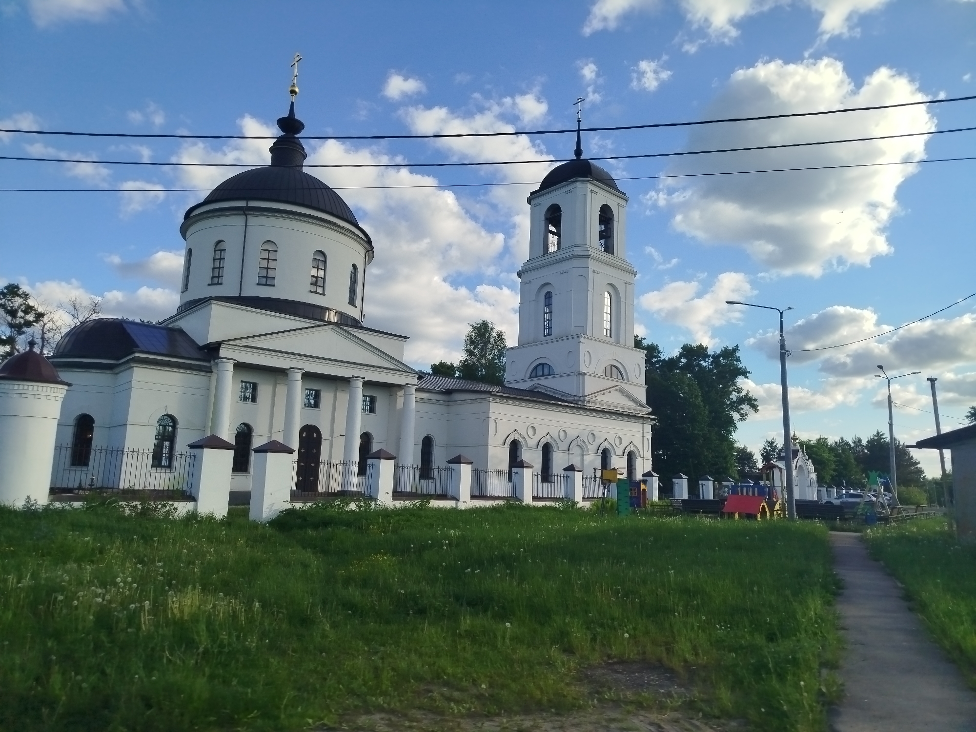 The image depicts the Church of the Nativity of the Theotokos in the village of Borovkovo, Russia. The church is white with dark domes topped by crosses. It has a large main structure with a dome and a separate bell tower. The church is surrounded by a fence with white posts. In front of the church is a grassy area. To the right, there is a sidewalk, with a playground and a small white chapel visible in the background. The sky is blue with scattered clouds. Power lines are visible running across the sky in the background.
