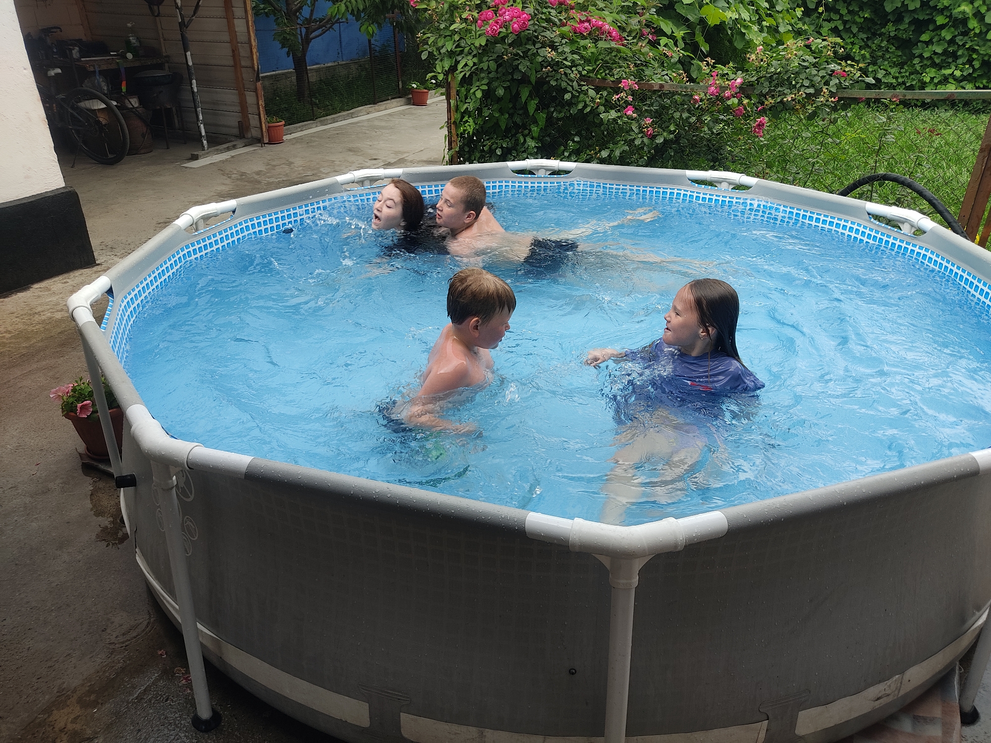 In this medium shot, four people are enjoying a refreshing dip in a hexagonal above-ground swimming pool filled with clear blue water. The scene appears to be set in a backyard on a warm, possibly overcast day, given the diffused natural light and the presence of lush green foliage and blooming pink flowers in the background.
The subjects include two adults and two children. One adult, a male, is lying on his back in the water, with his head resting on the pool's edge. Next to him, a young girl is looking up and to the side, with water splashing around her face. A young boy is in the foreground, facing left, with his arms outstretched in the water. Another young girl, wearing a blue swimsuit, is also in the pool, looking to the left with a slight smile. The pool itself has a blue tiled interior and a grey outer wall, supported by white piping and metal legs.
In the background, to the left of the pool, there's a glimpse of a covered area with a bicycle and some stored items, suggesting a residential setting. The overall mood is one of playful relaxation and enjoyment of a summer day. The location is Shamalgan, Kazakhstan. There is no visible text in the image.
