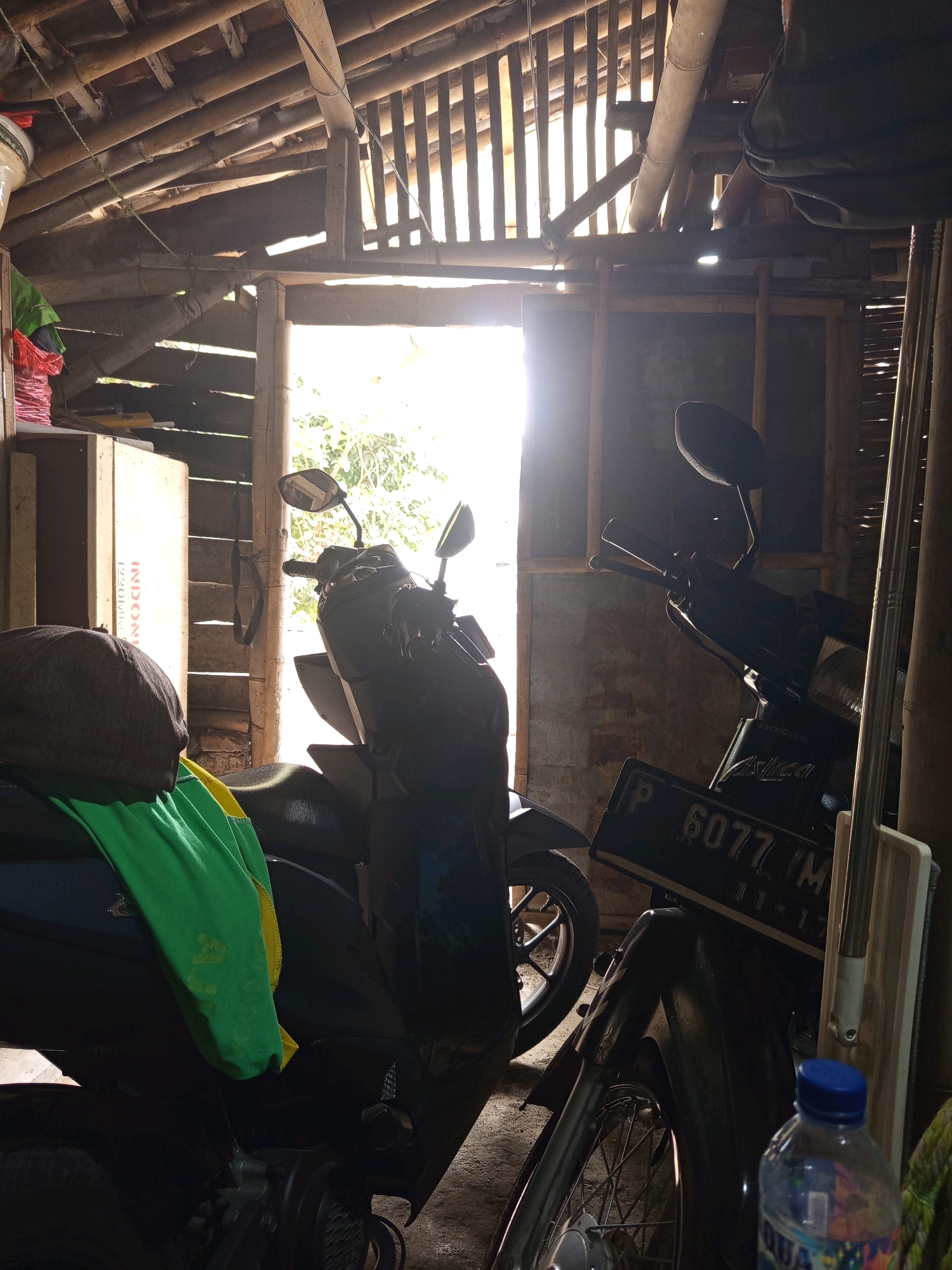 The image depicts the interior of a rustic structure, possibly a barn or shed, in Rowo Indah, Indonesia. The structure features a roof made of bamboo poles and battens, with light filtering through gaps, creating a dappled effect. In the foreground, a dark-colored scooter is partially visible, with a green and yellow cloth draped over its seat and handlebars. To the right, another scooter is more prominently featured, also dark-colored. This scooter displays a license plate with the characters "P 6077 VM". A clear plastic bottle with a blue cap, partially filled with water, is positioned in the lower right corner of the frame. The background shows a doorway with bright daylight streaming in, revealing foliage and trees outside. The walls appear to be made of rough, possibly unfinished materials. The overall impression is one of a humble, rural storage space. There are no people visible in the image.